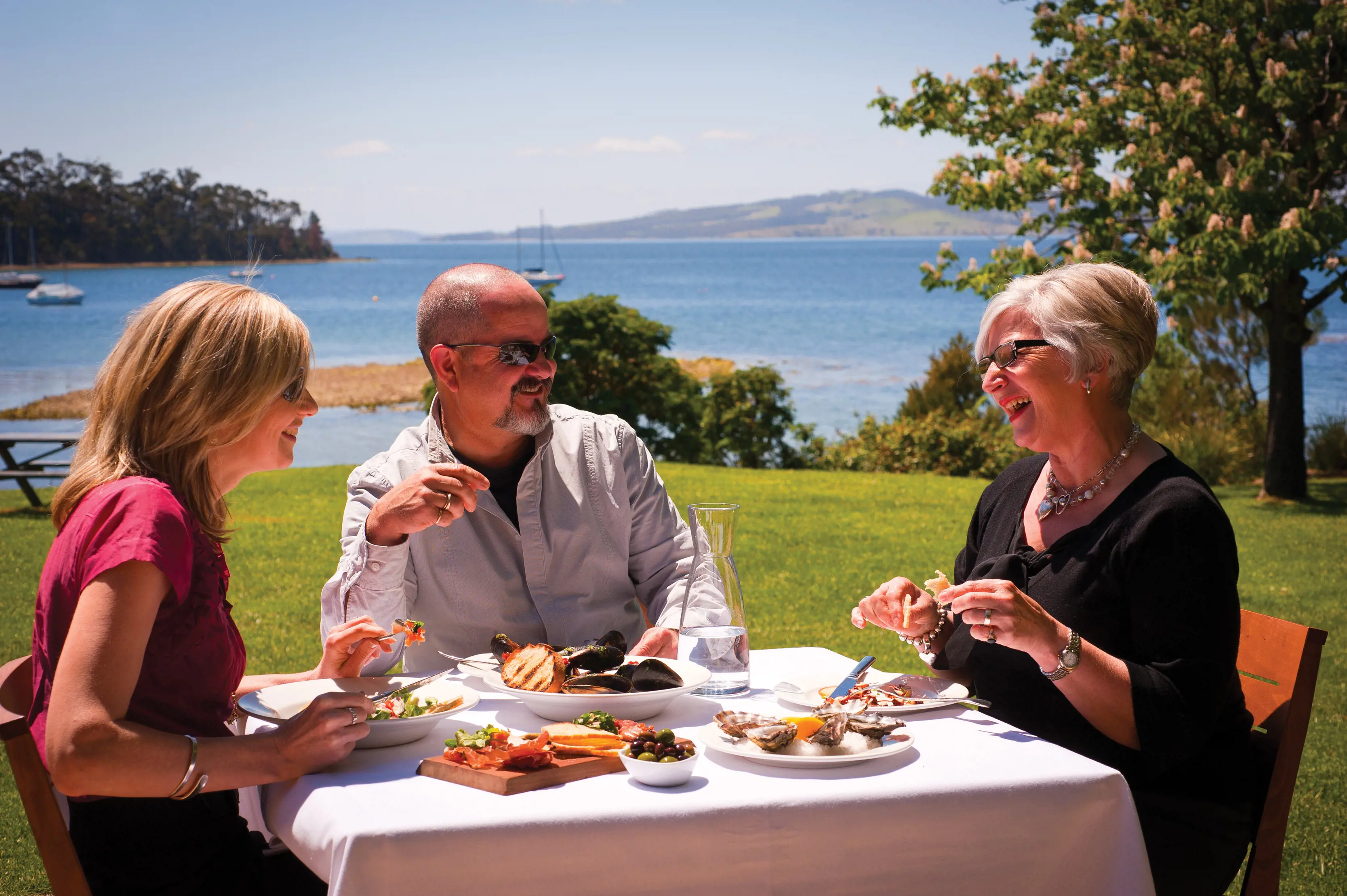 A group of people enjoy food sat outdoors on the grass at Stackings Restaurant, Peppermint Bay. Water and mountains in the backdrop.