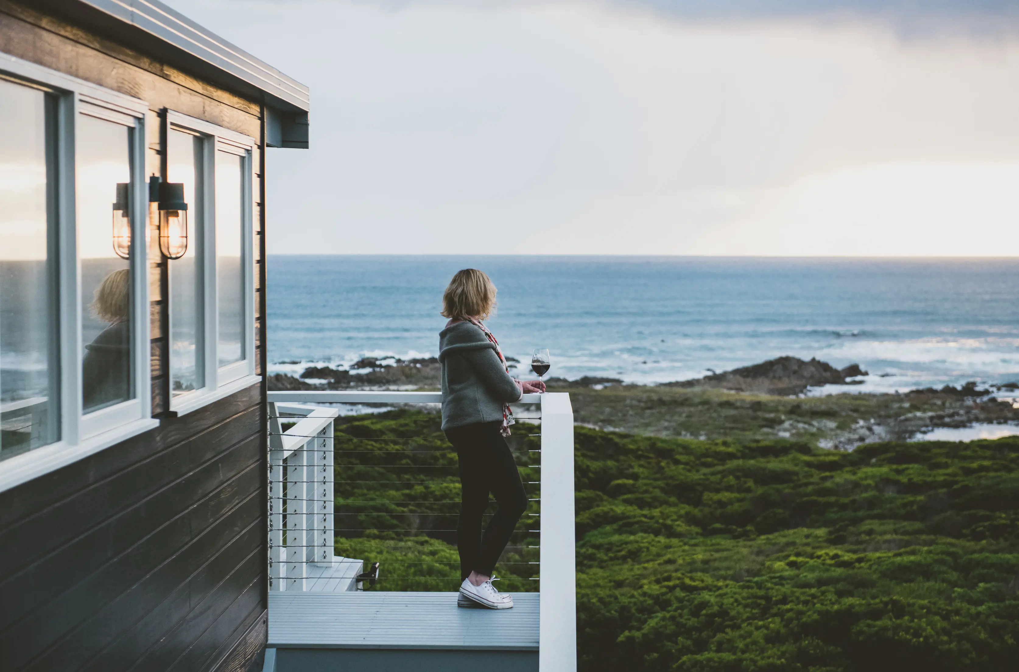 A woman leans on the balcony outside the Taraki Lodge, looking out the ocean and landscape.