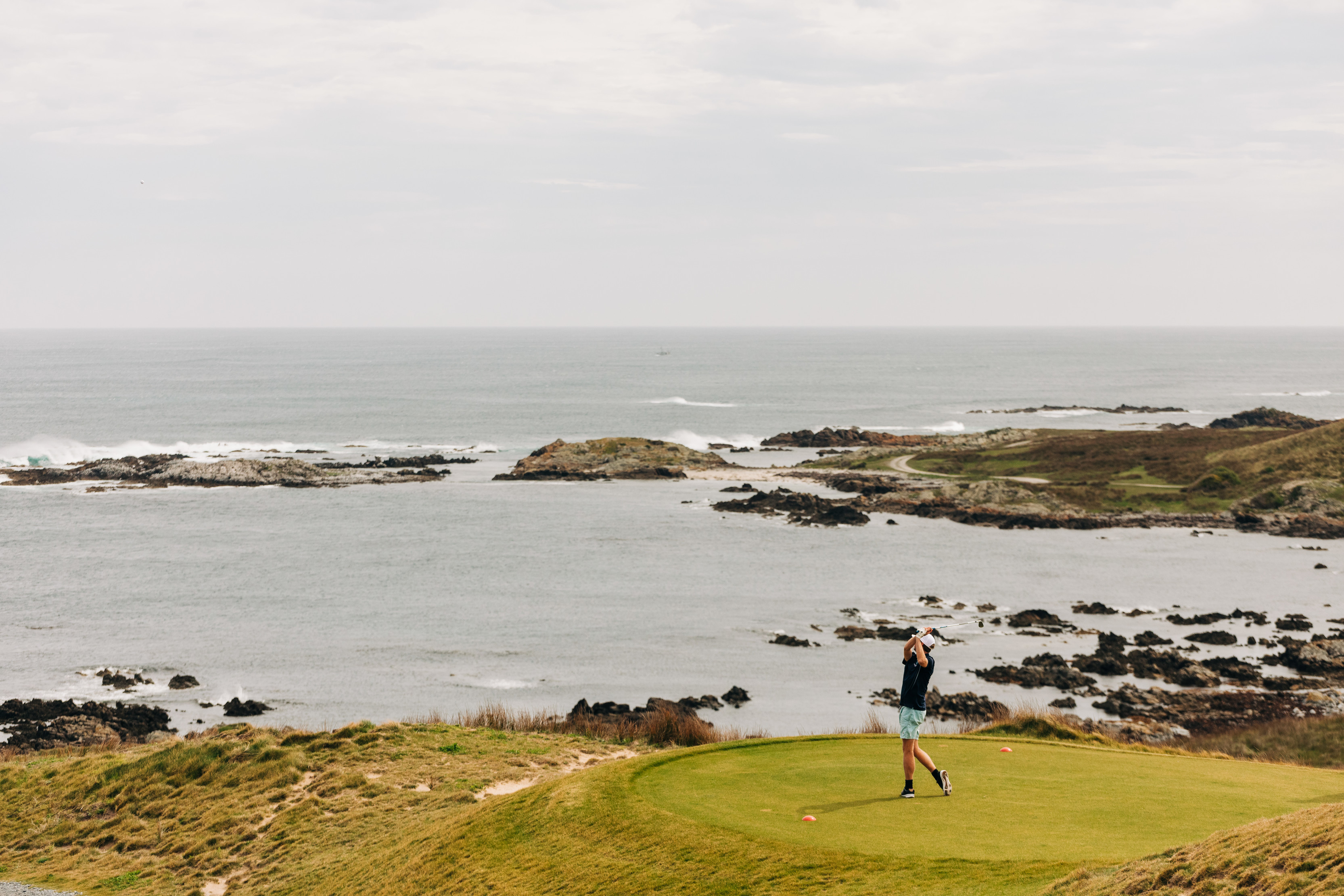 Person swinging the golf club on the turf, surrounded by incredible scenes of rock, hills and ocean, beside the Great Southern Ocean.