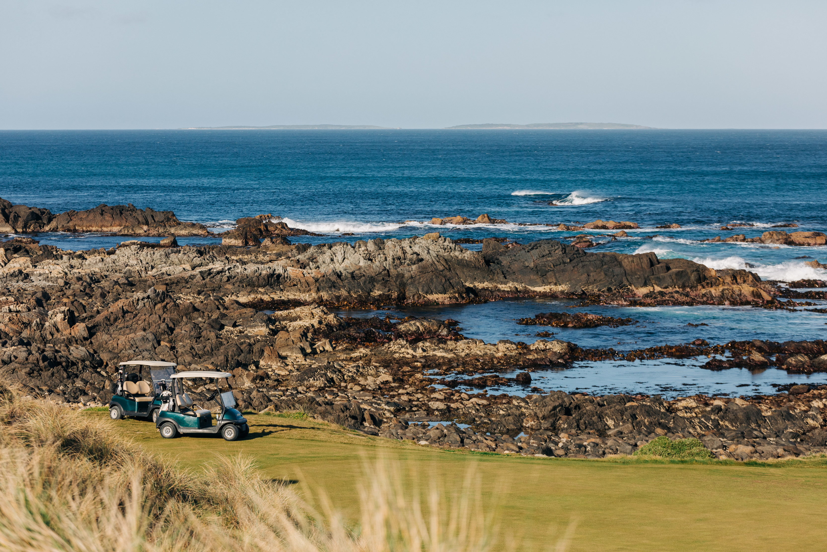 Two gold buggies parked on the golfing green, right by the rocks and ocean, located at Cape Wickham Golf Course. 