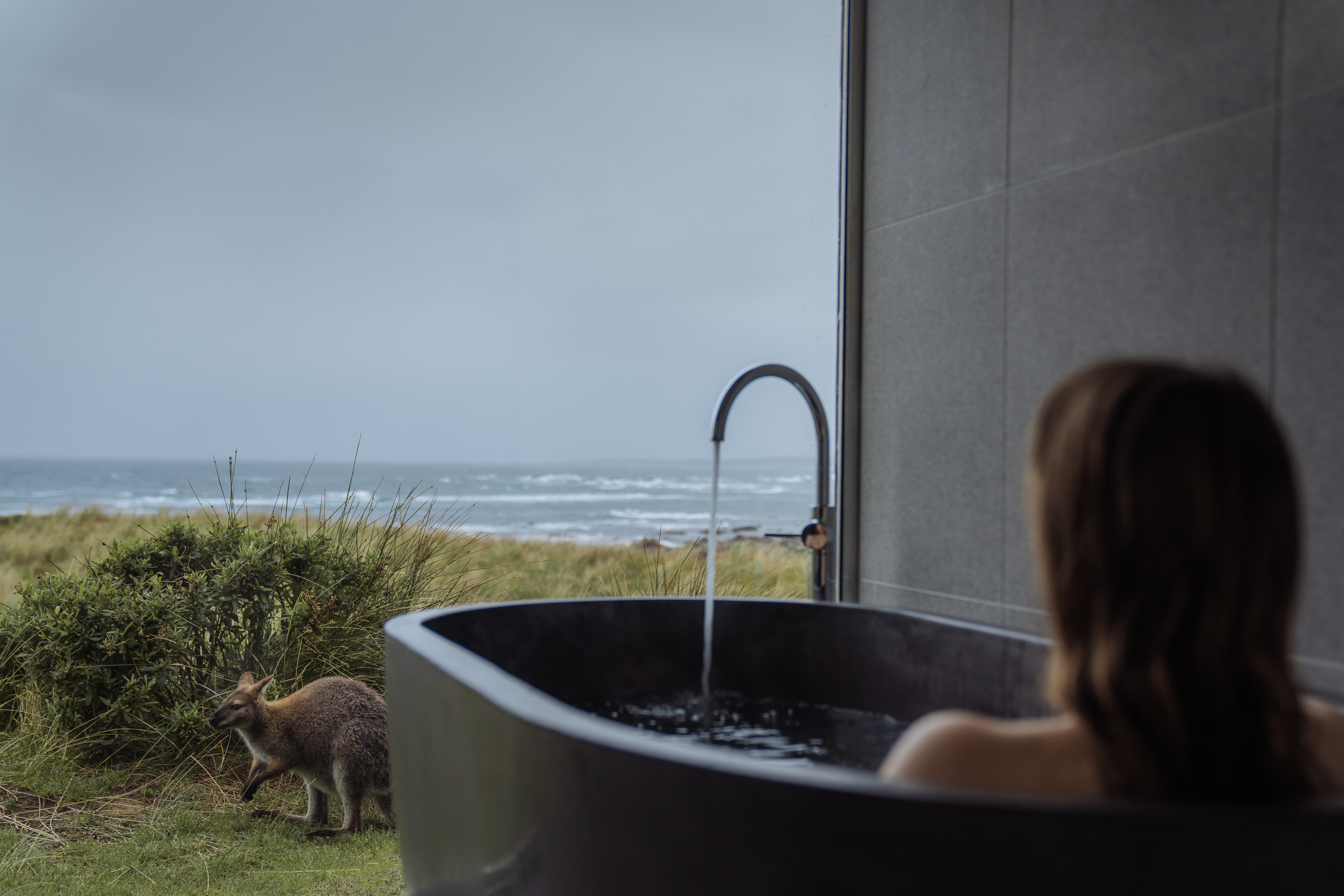 Woman relaxing in the outdoor bath with a kangaroo nearby, located at Kittawa Lodge.