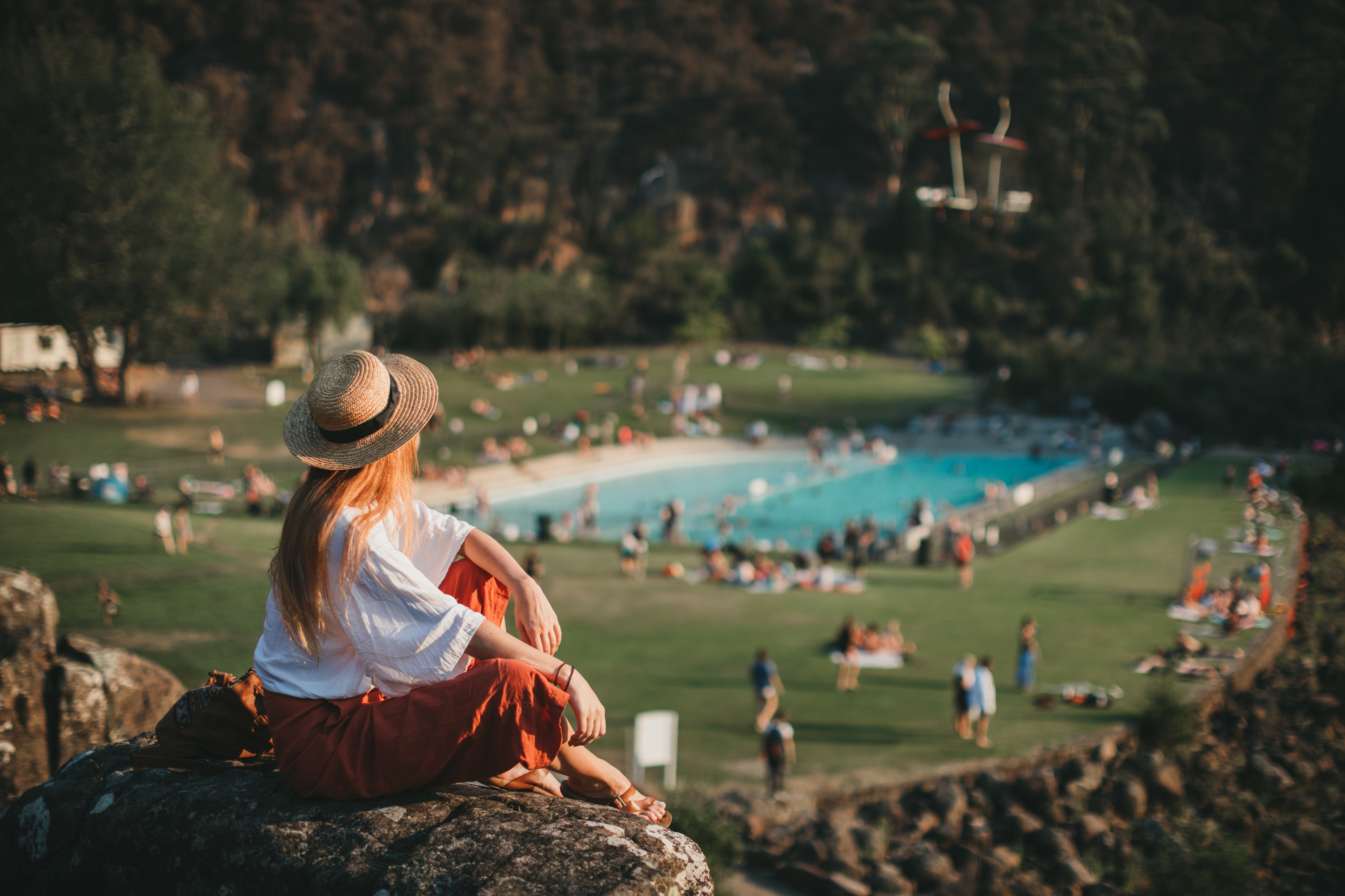 A woman sits on a rock looking out at crowds enjoying the swimming pool at Cataract Gorge. A pool surrounded by rainforest.