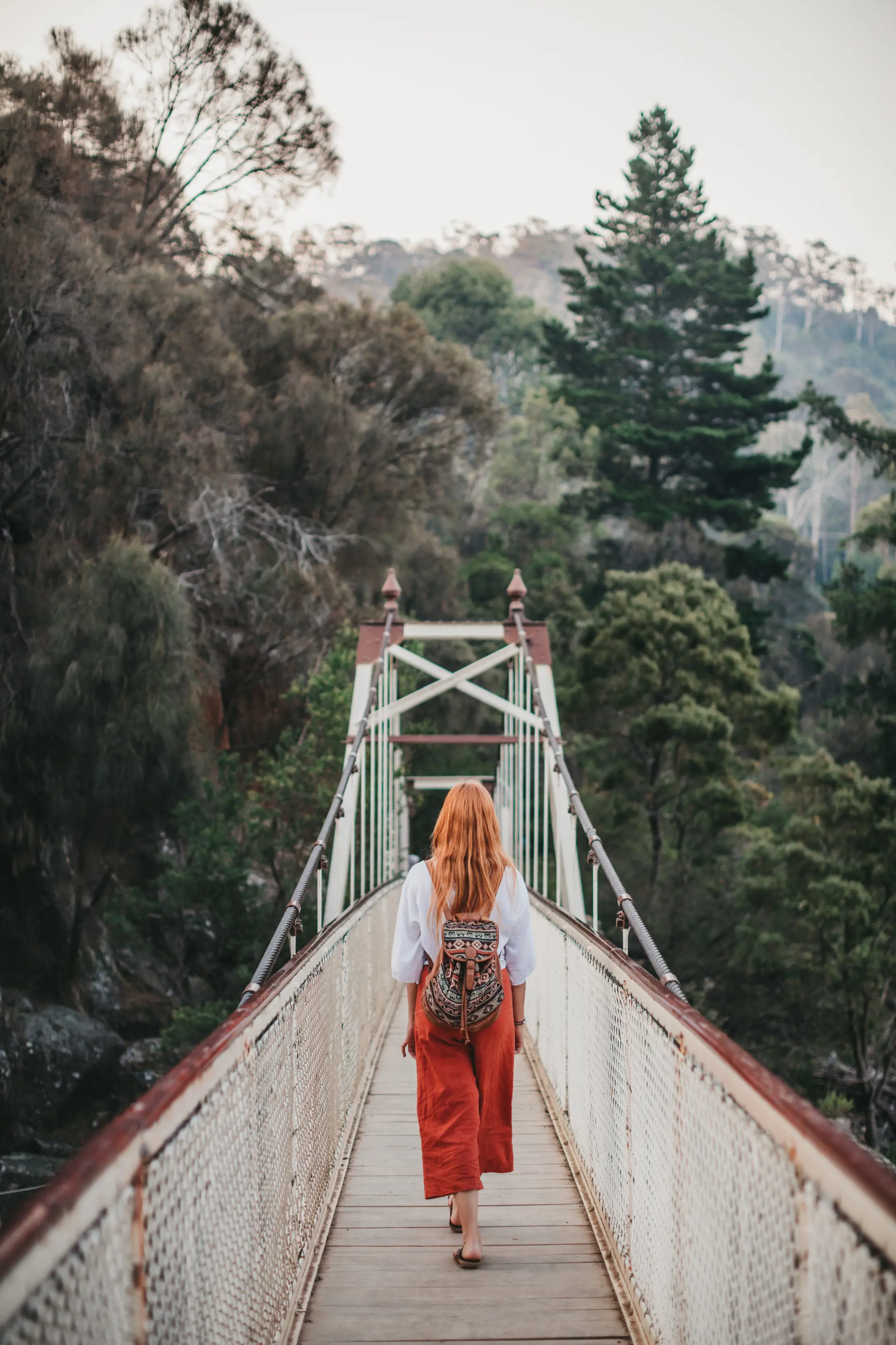 A woman with red hair walks across the Suspension bridge at Cataract Gorge Reserve. There is mist over the surrounding rainforest.