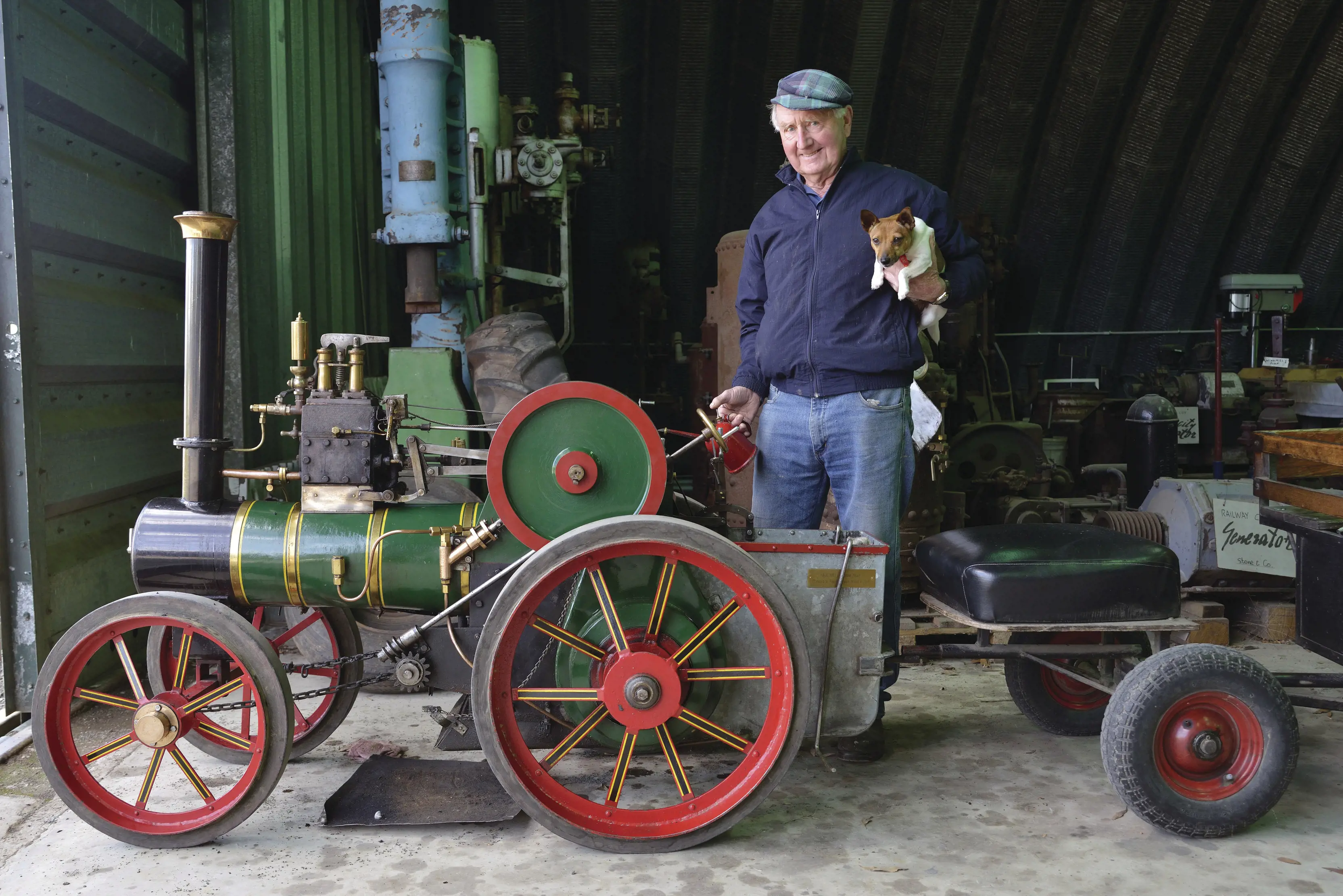 Man, standing behind the head of an old fashioned, oversized toy train head. Dog in one hand and oil can in the other, at the Pearns Steam World