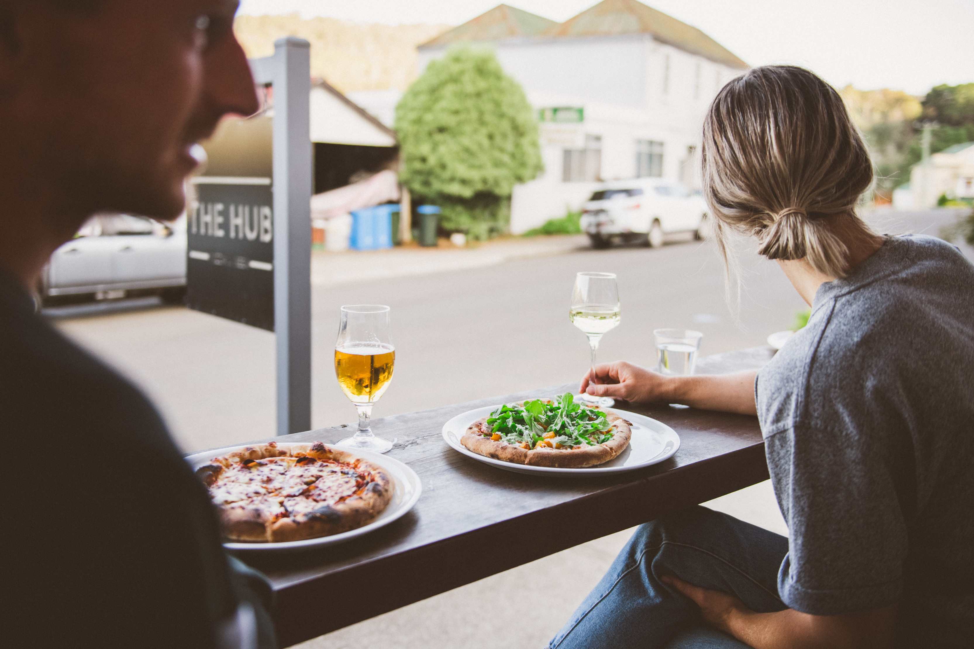 A couple enjoy pizza, wine and beer whilst sitting on a bench looking out at the street at the front of The Hub - Pizza and Beer.