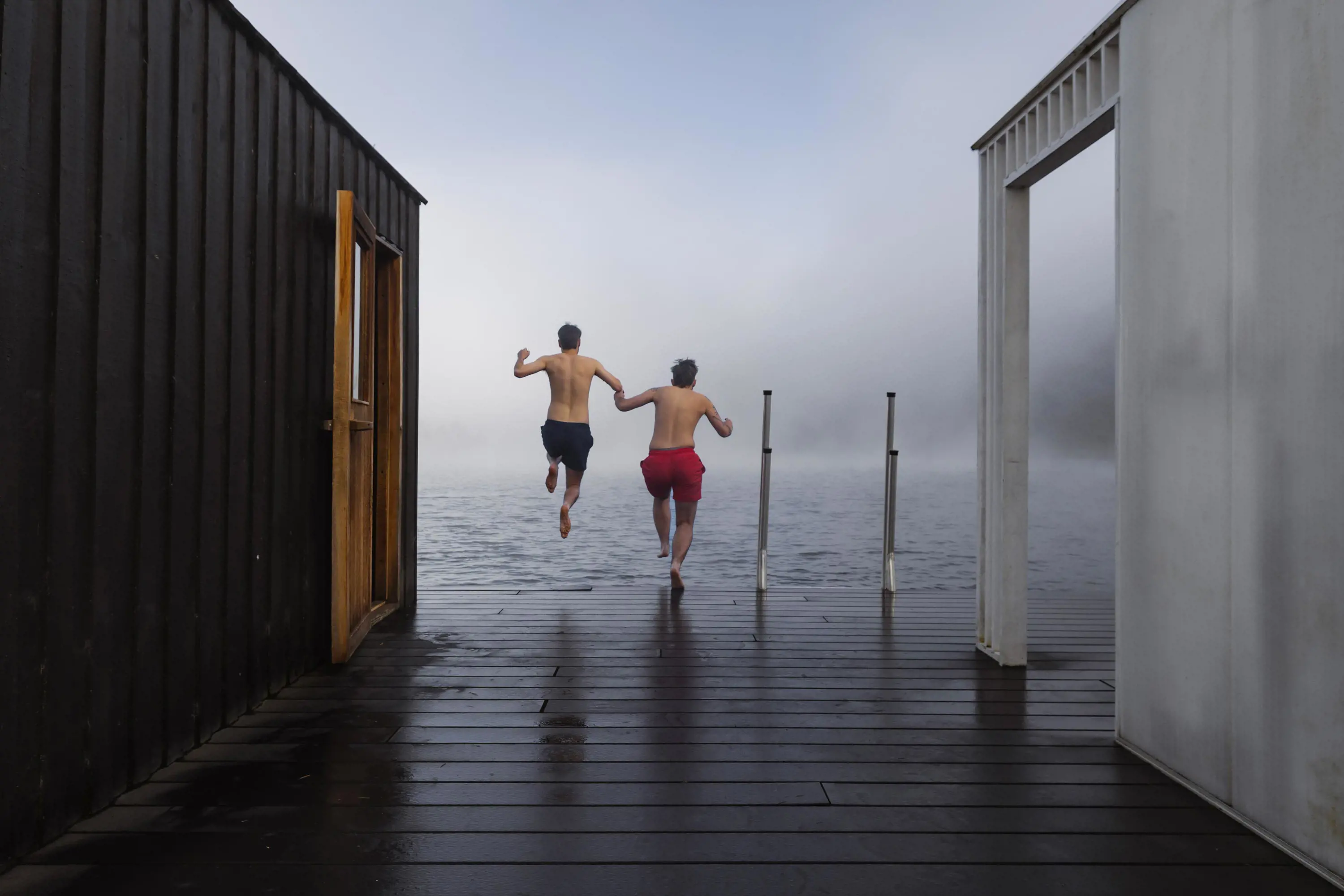 To young men holding hands jump into the cold waters of a lake from the decking in front of a sauna.