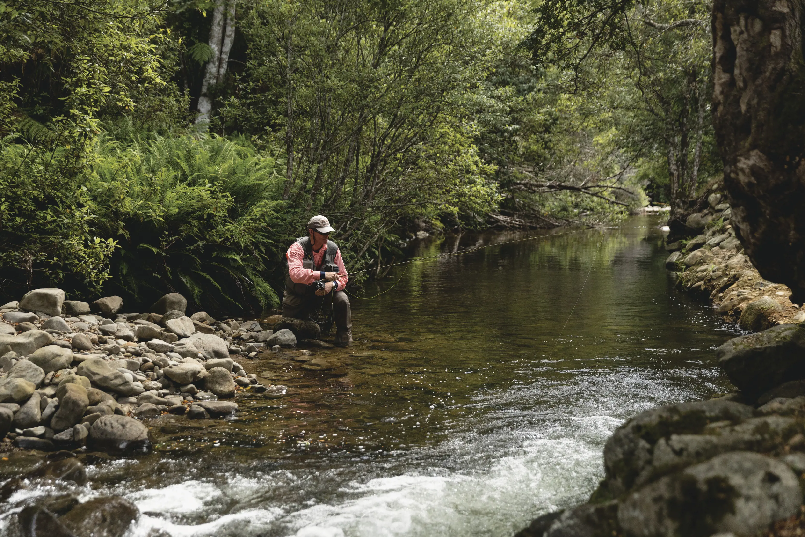 A man in a pink shirt crouches in shallow water whilst fly fishing on the Liffey River.