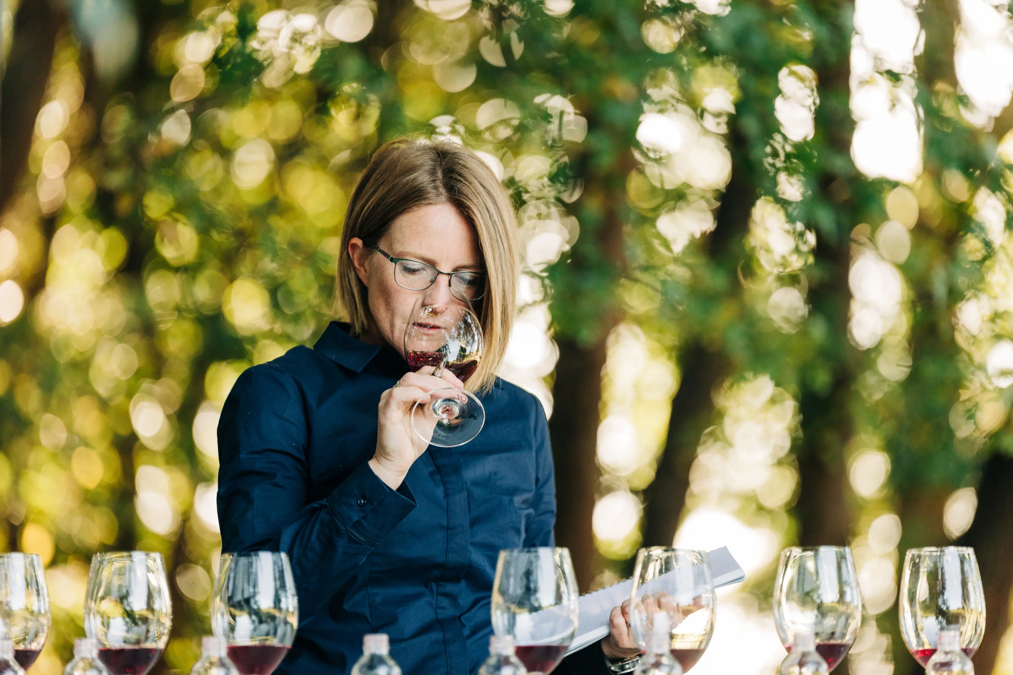 A woman enjoys wine tasting outdoors at Holm Oak Vineyards. She holds a notepad in one hand and wine glass to her nose in the other.