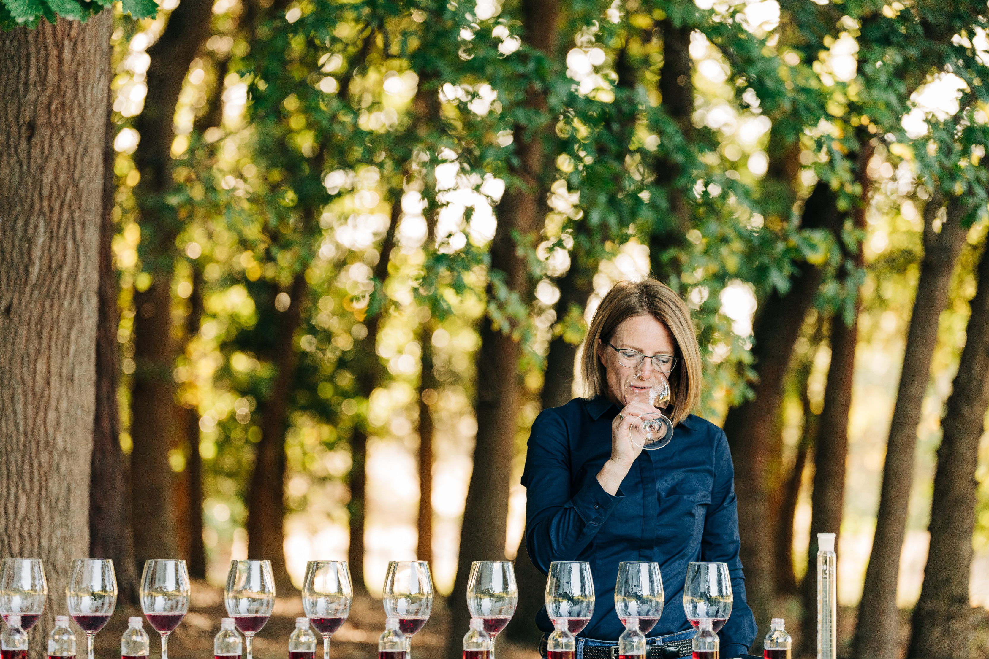 10 glasses of red wine are placed in a row outdoors at Holm Oak Vineyards wine tasting. A woman tastes wine from one glass.