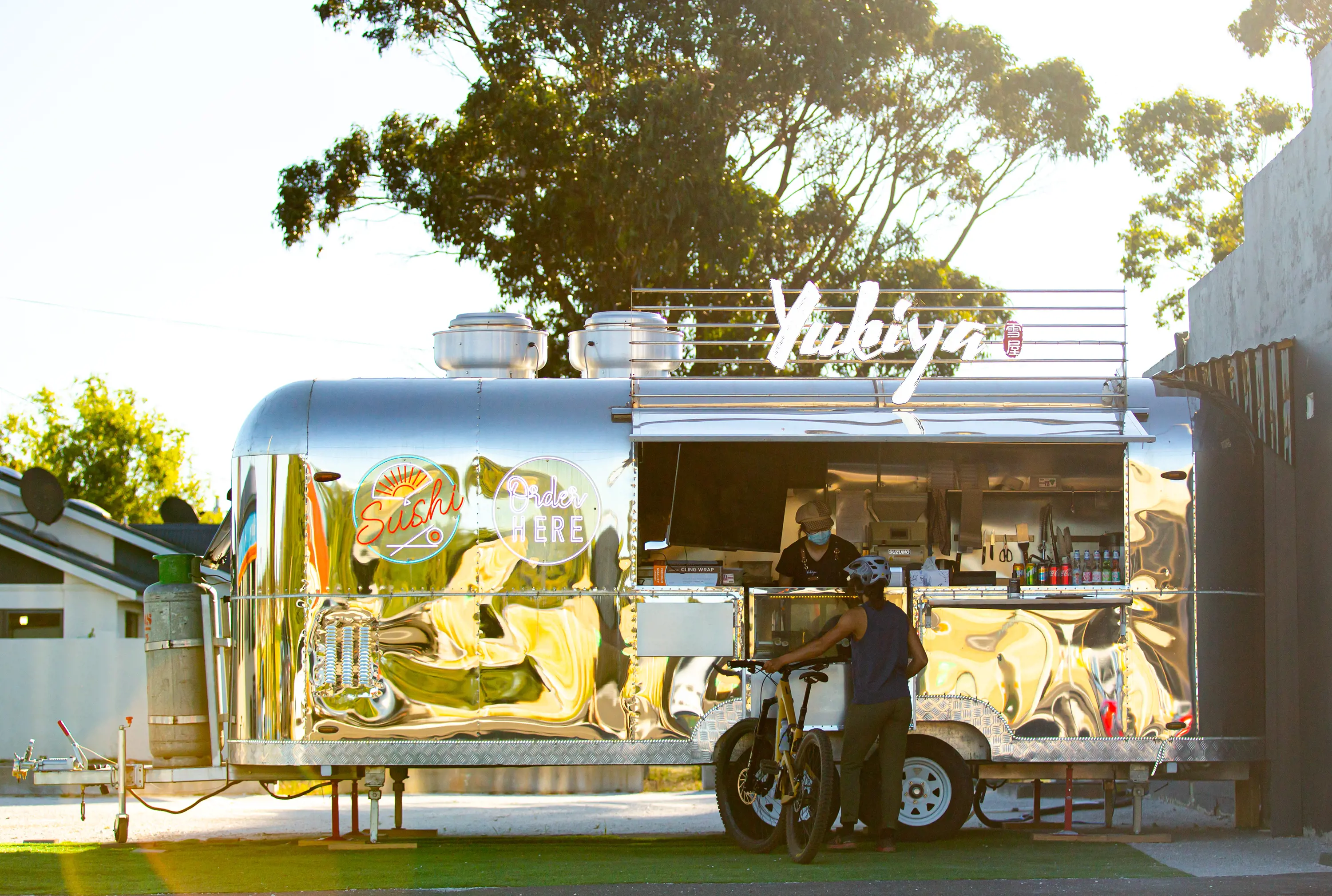 A customer with a bike stands in front of a polished aluminum caravan that is serving sushi.