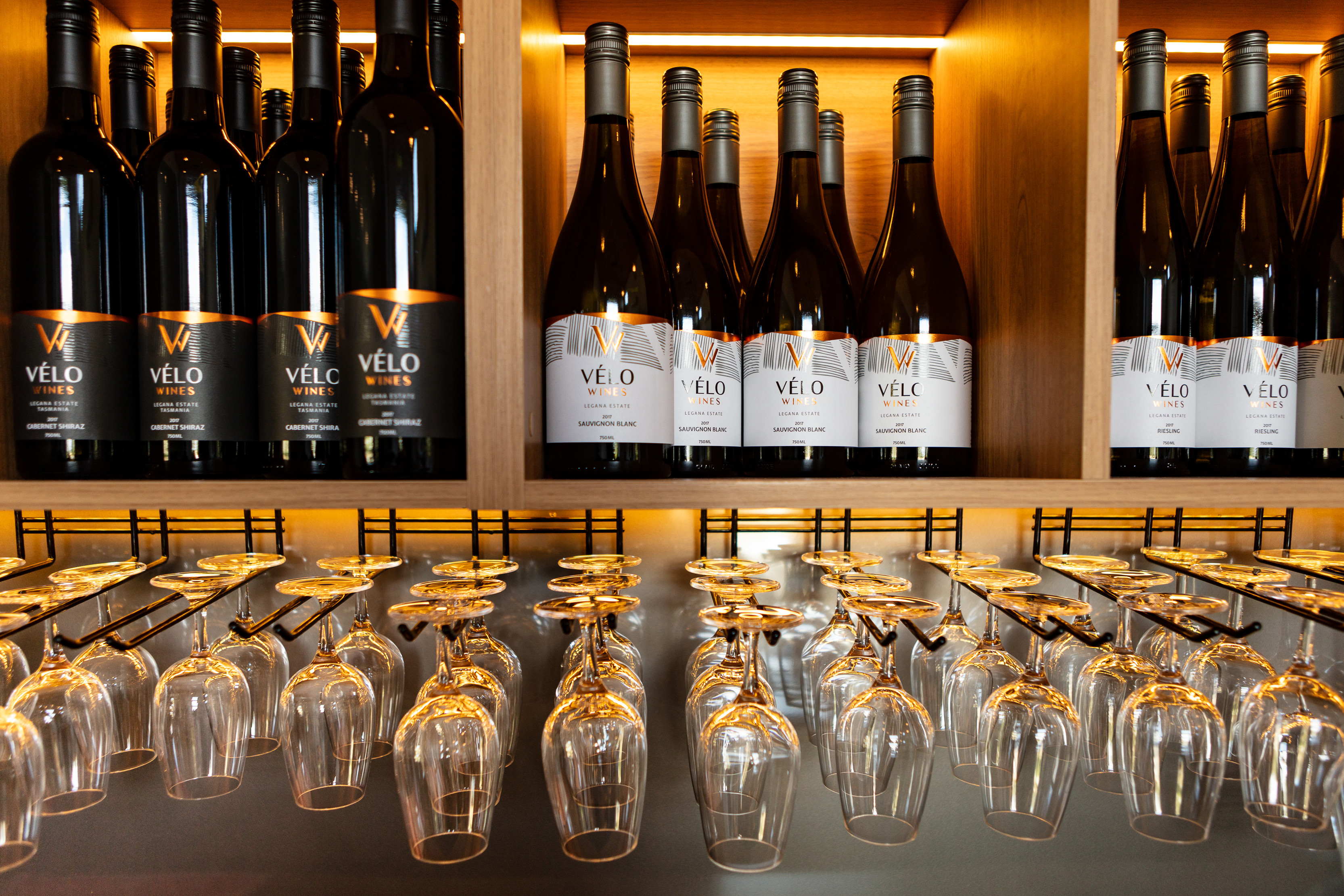 A variety of Velo wines sit above in shelves as wine glasses hang underneath.