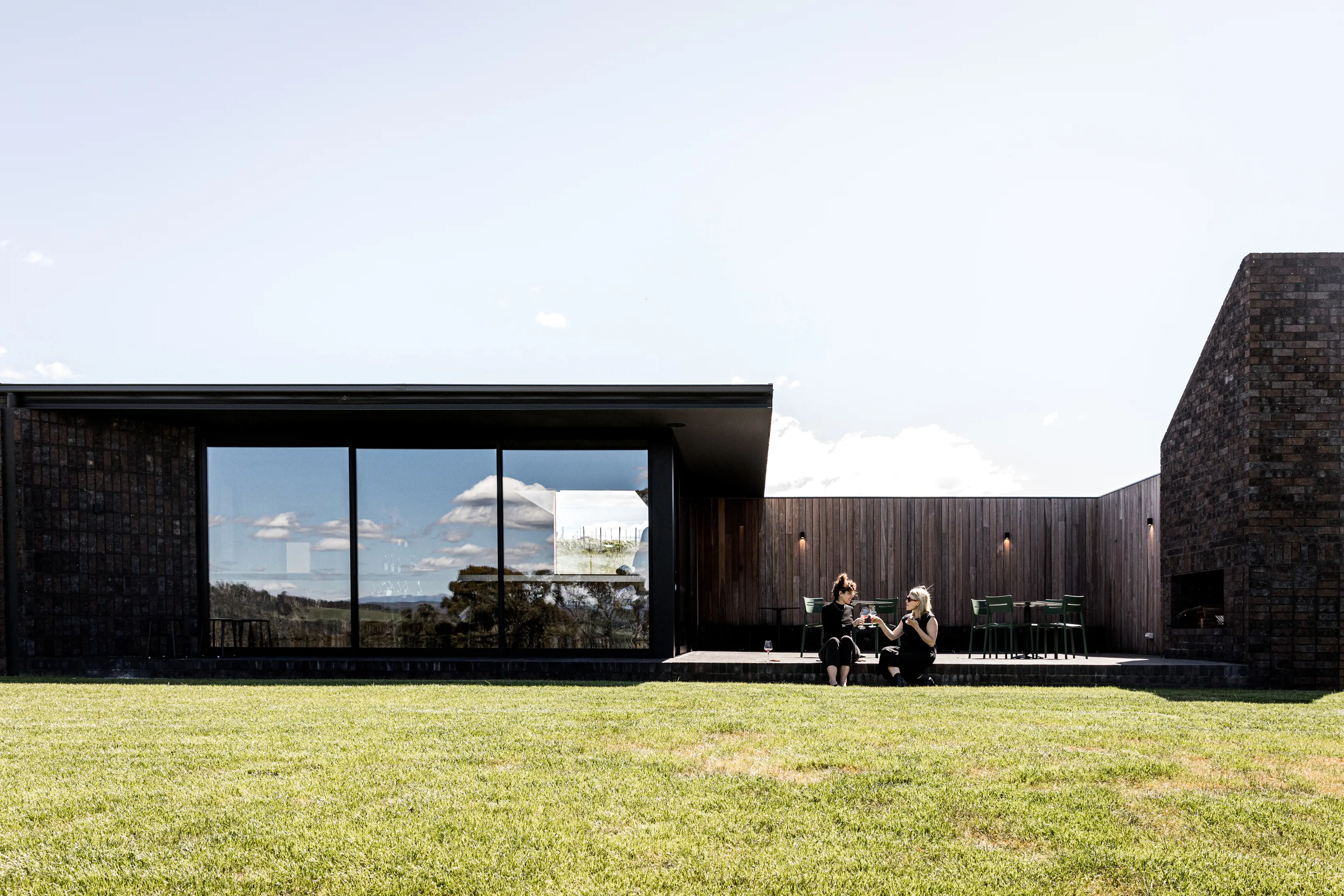 Two friends enjoy a drink outside the modern grey exterior of Stoney Rise Wine Company.