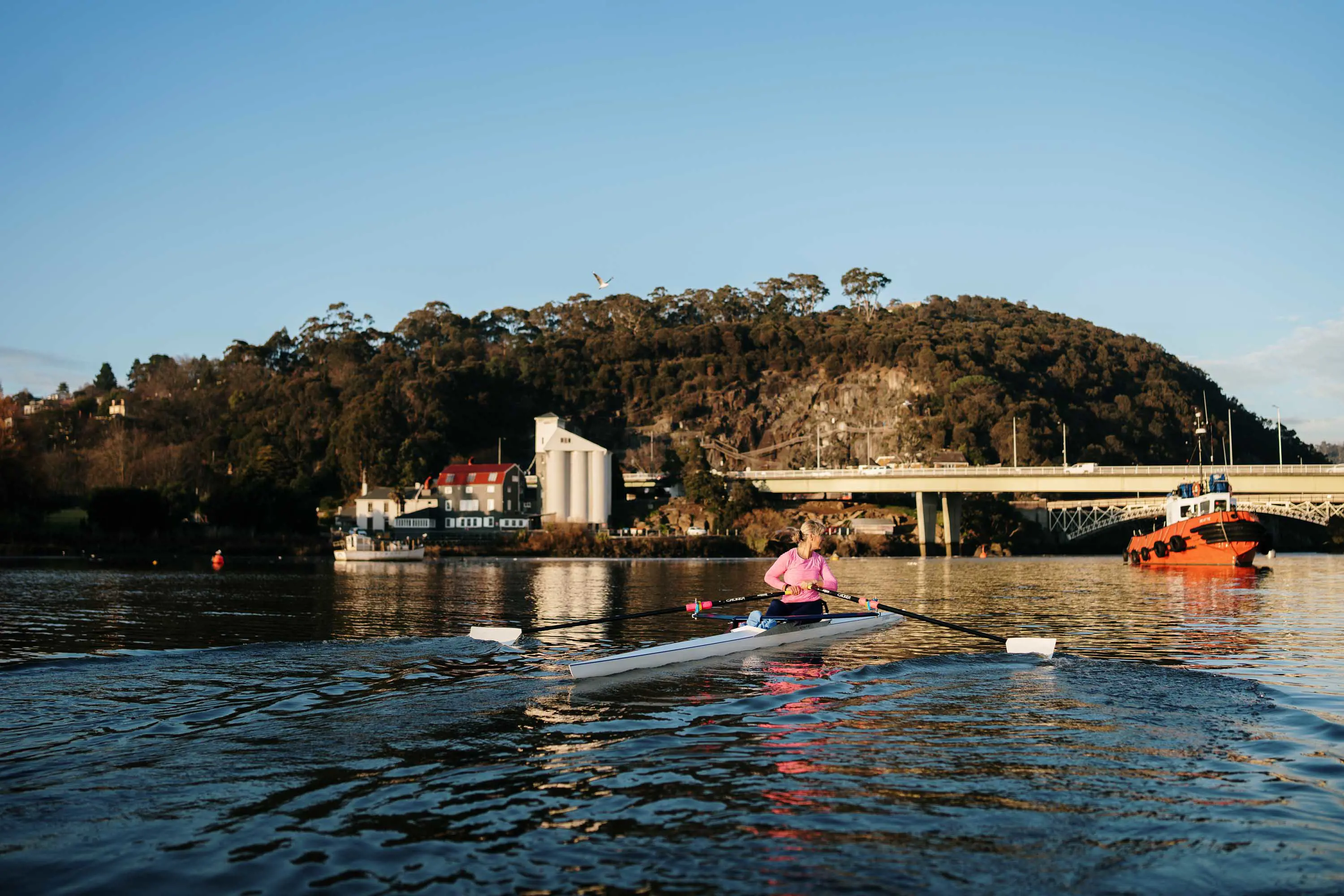A woman rows a canoe towards a long bridge in fine weather.
