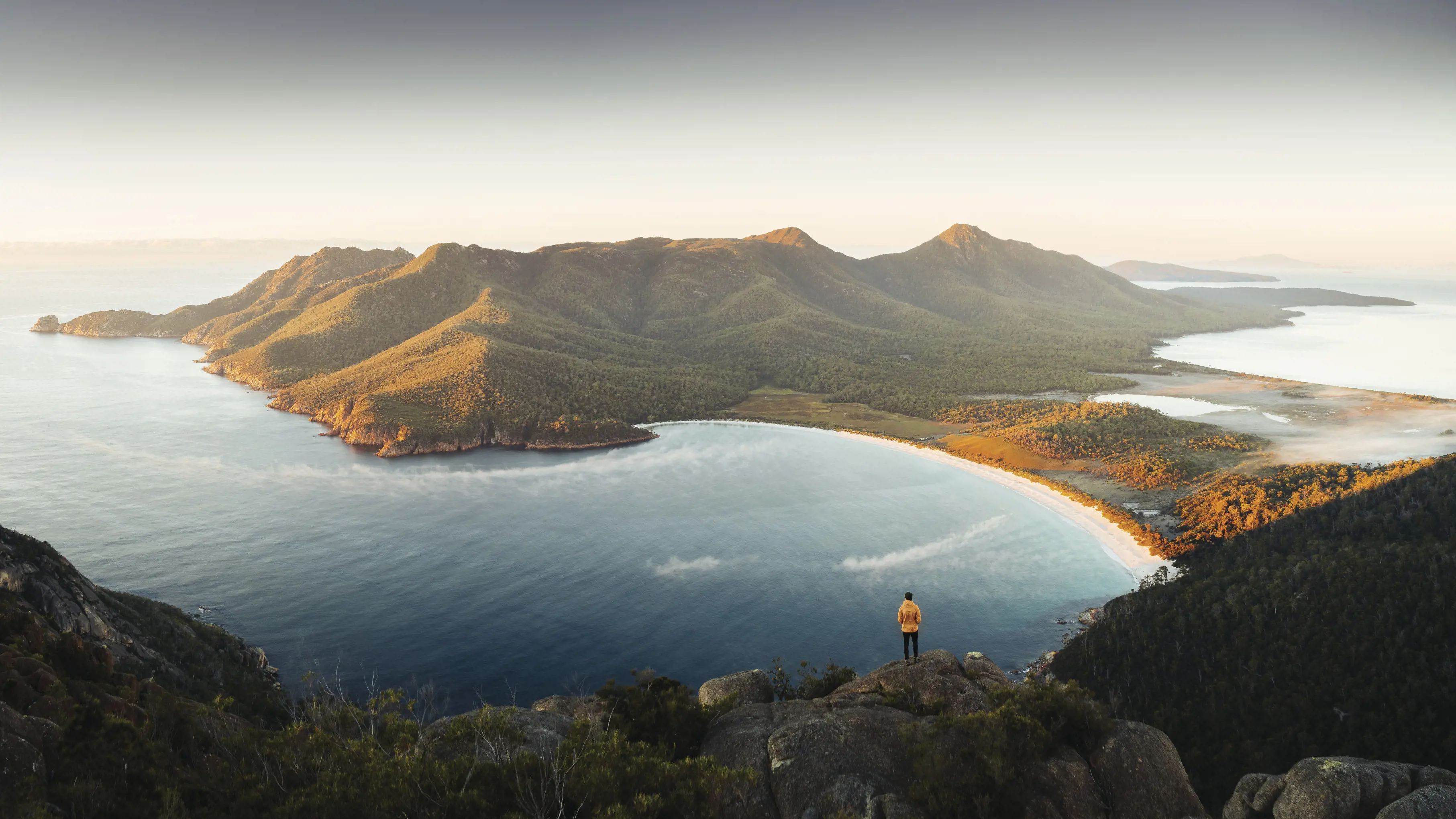 Aerial shot of Wineglass Bay, Freycinet National Park. A man standing on the cliff, looking out onto the water. 