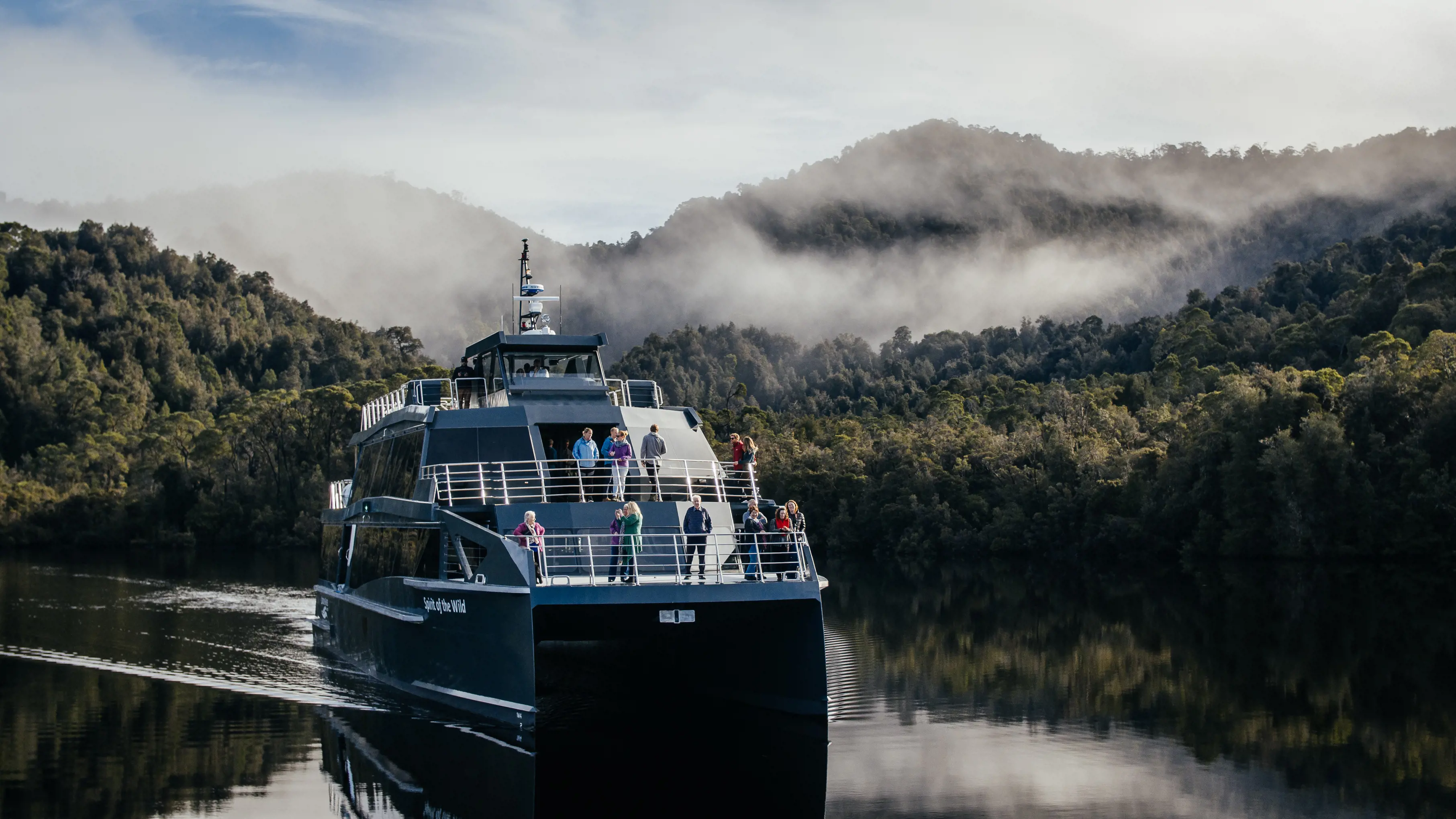Spectacular image of Gordon River Cruises with people at the front of the boat, piloting through Gordon River, surrounded by lush forest.