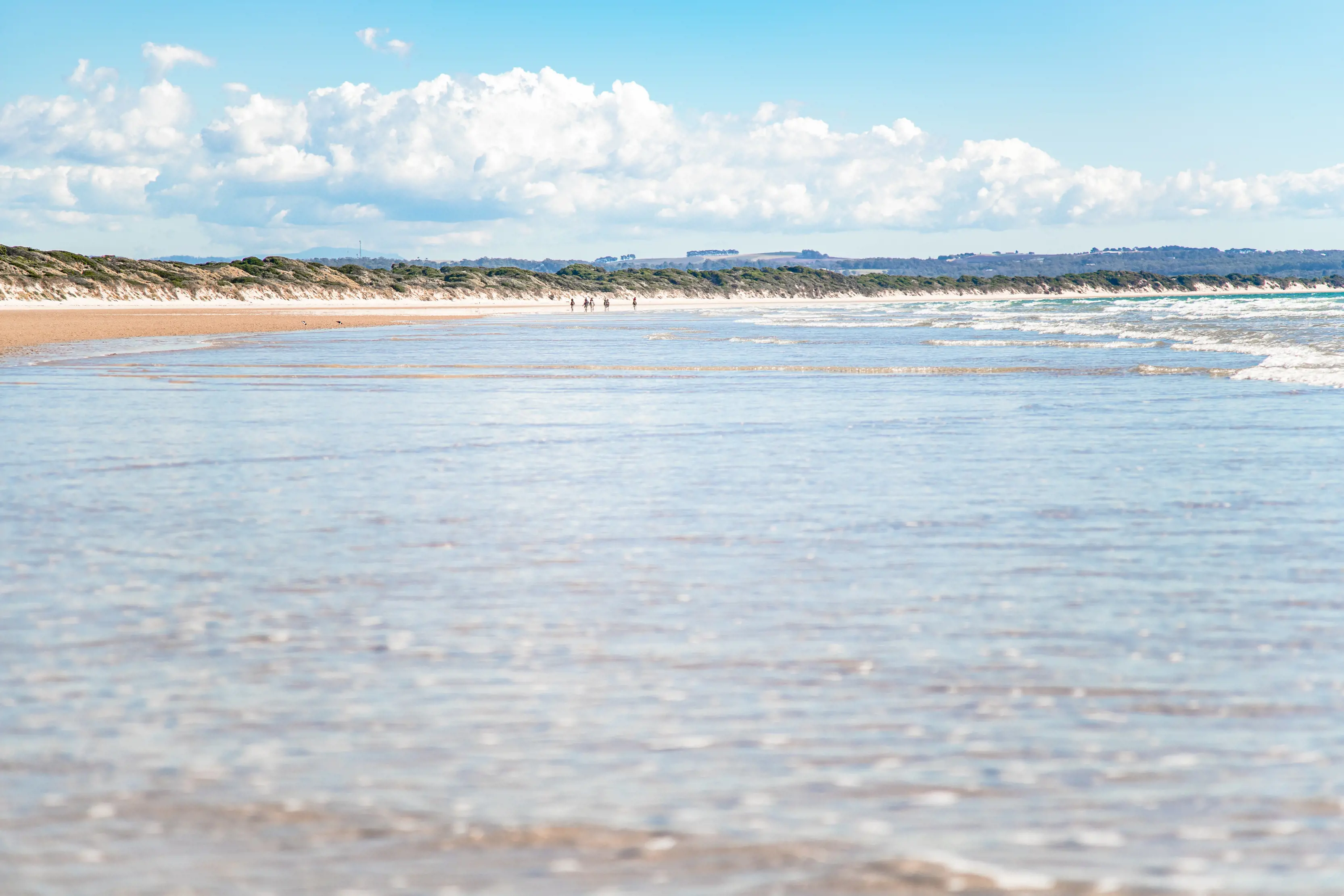 Low image taken of the shoreline of Baker's Beach, with crystal clear water and people in the distance on the sand, walking along the beach.