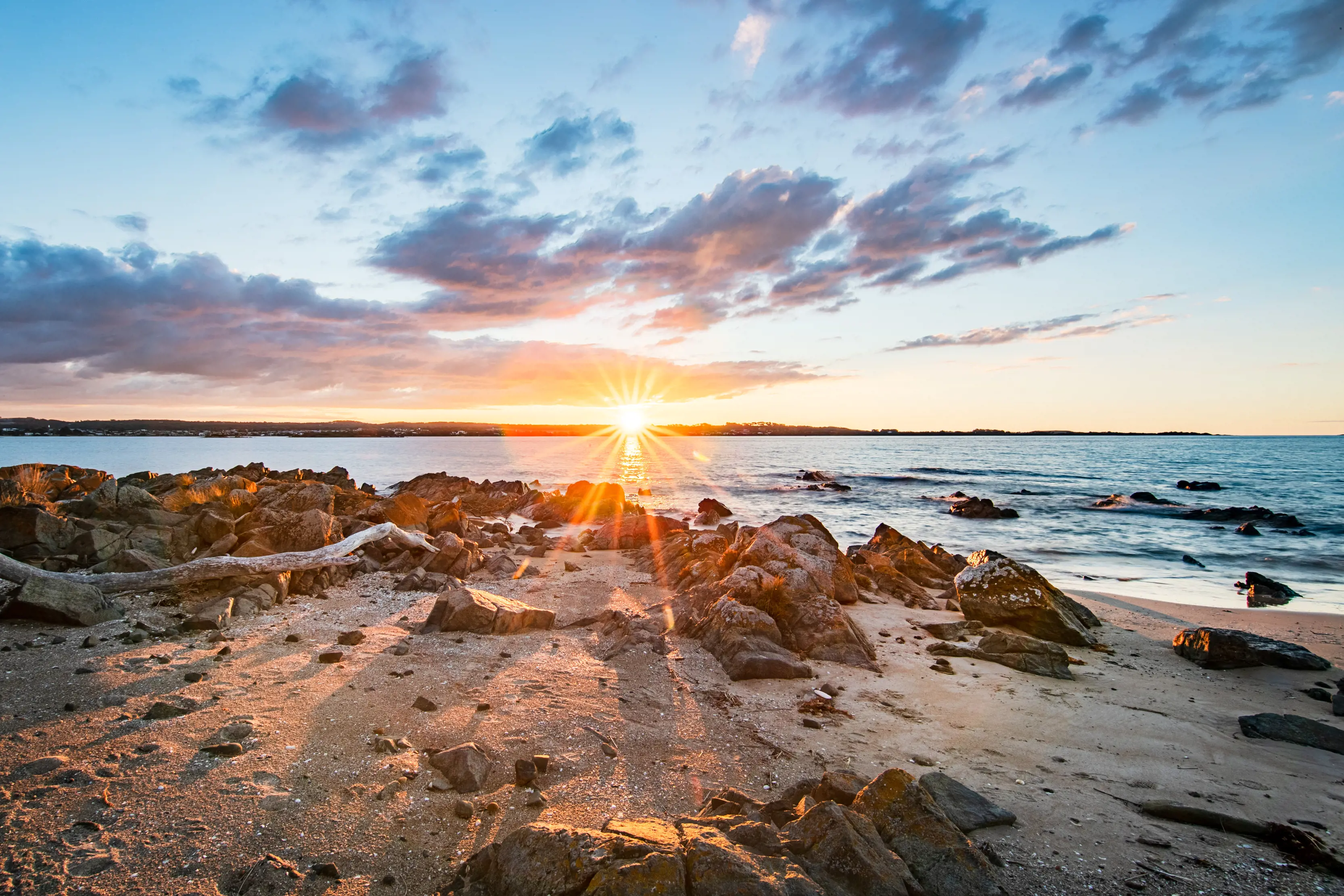 Wide angle image taken during a vibrant sunrise, sand rocks fill the foreground, running towards the ocean at Griffiths Point.