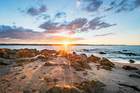 Wide angle image taken during a vibrant sunrise, sand rocks fill the foreground, running towards the ocean at Griffiths Point.