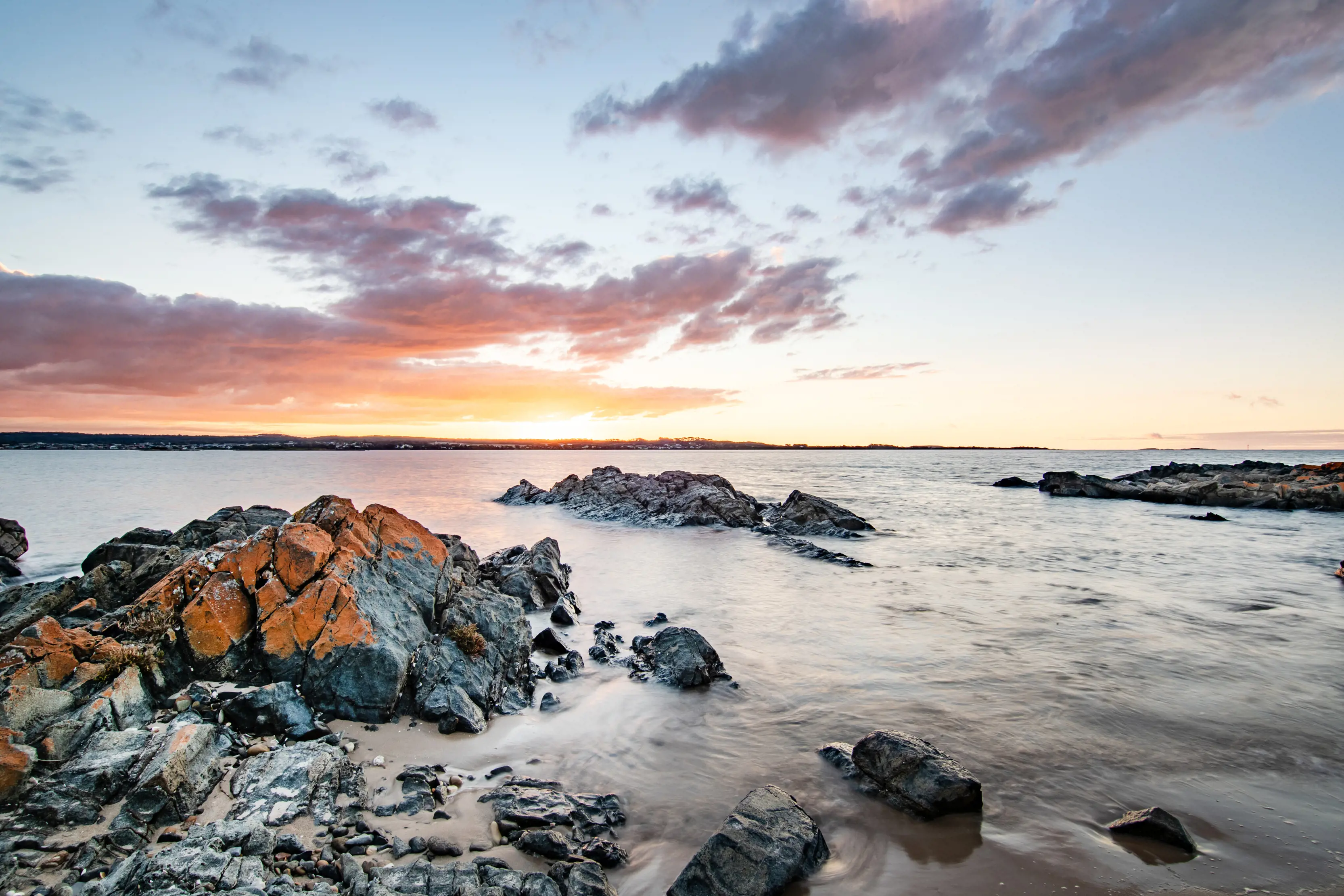 Wide angle image taken during a vibrant sunrise, large rocks in the sand fill the bottom left corner, running towards the ocean at Griffiths Point.