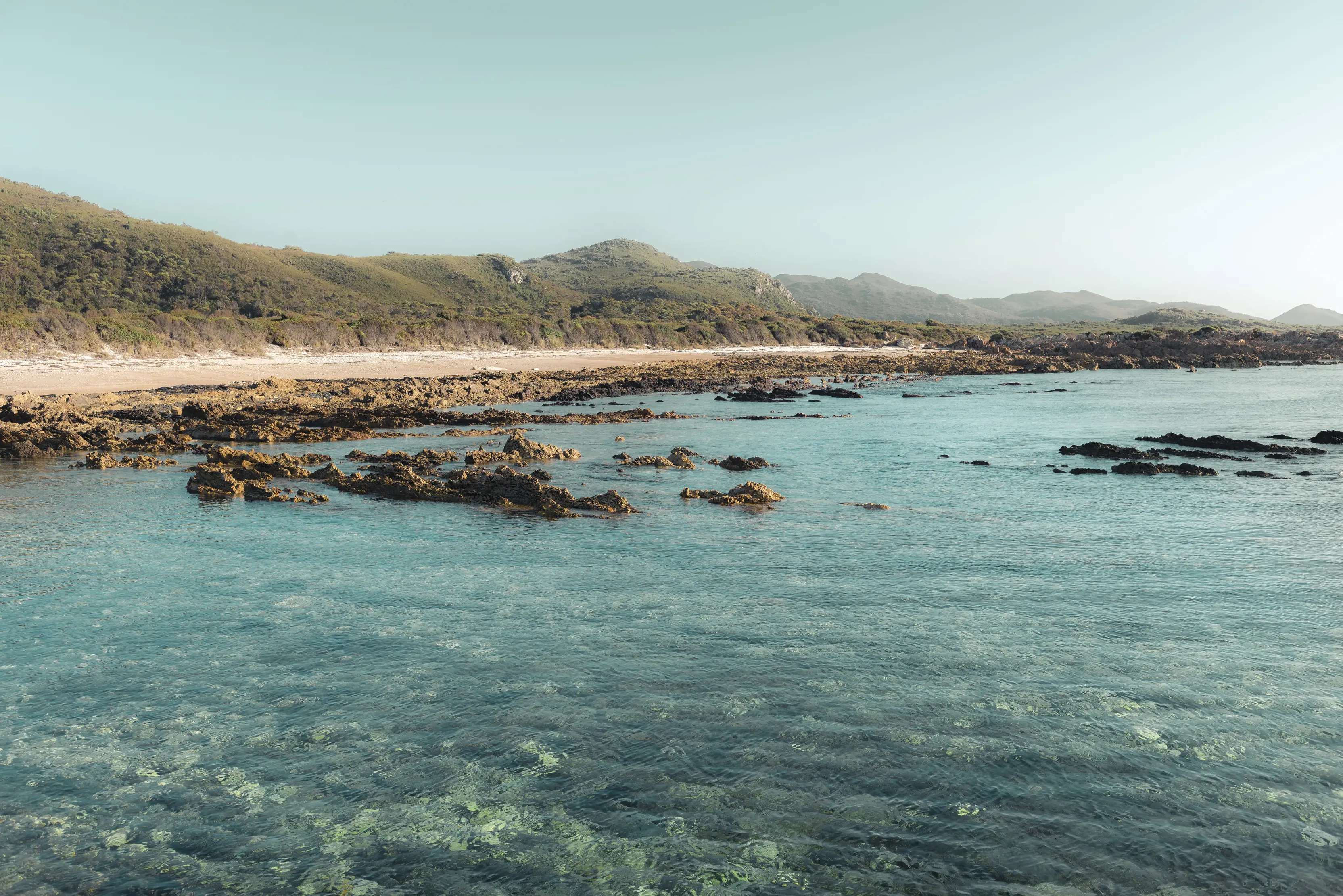 "Stunning image of Anniversary Bay. Crystal blue water fills the foreground with the shoreline running along in the background that meet with lush greenery. "