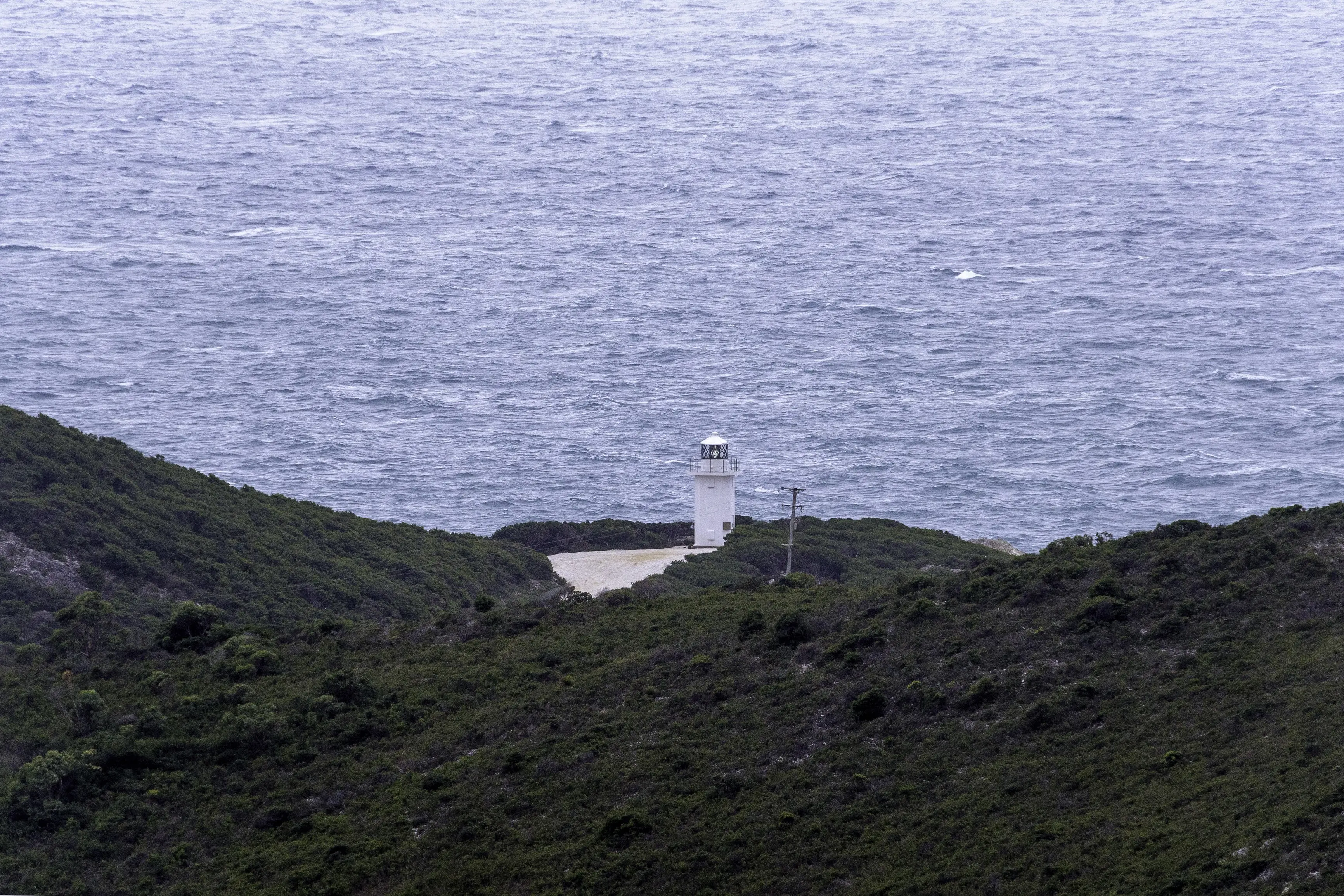 "Image of the Rocky Cape Lighthouse in the distance with large, rocky hills fill the foreground, on an overcast day. "