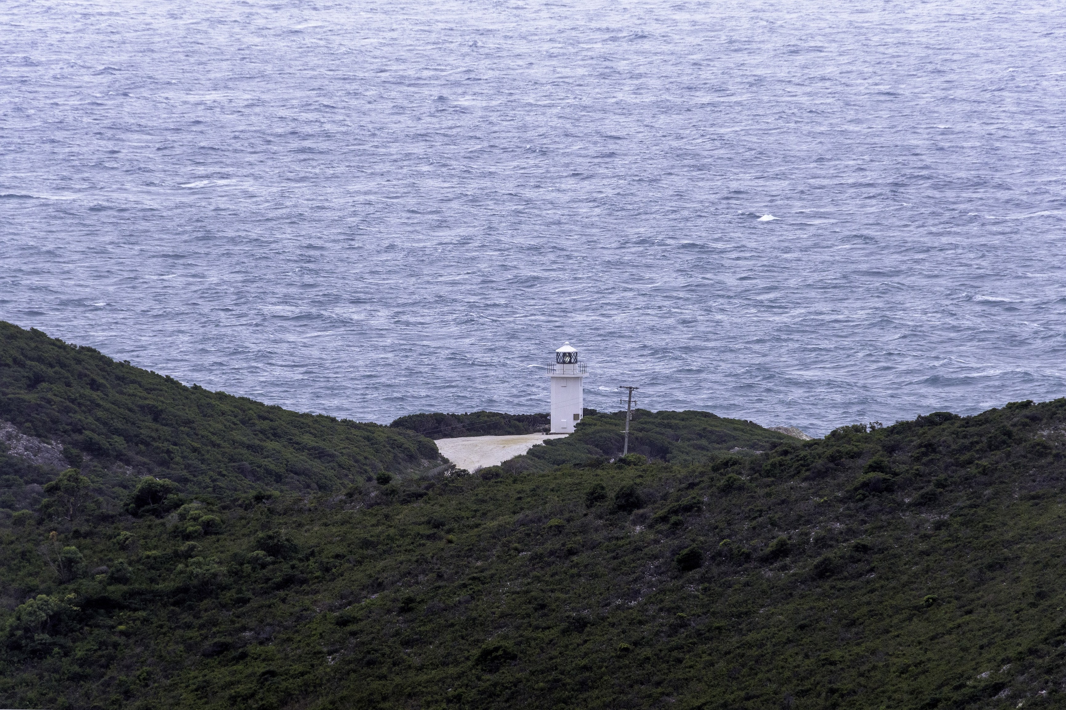 "Image of the Rocky Cape Lighthouse in the distance with large, rocky hills fill the foreground, on an overcast day. "