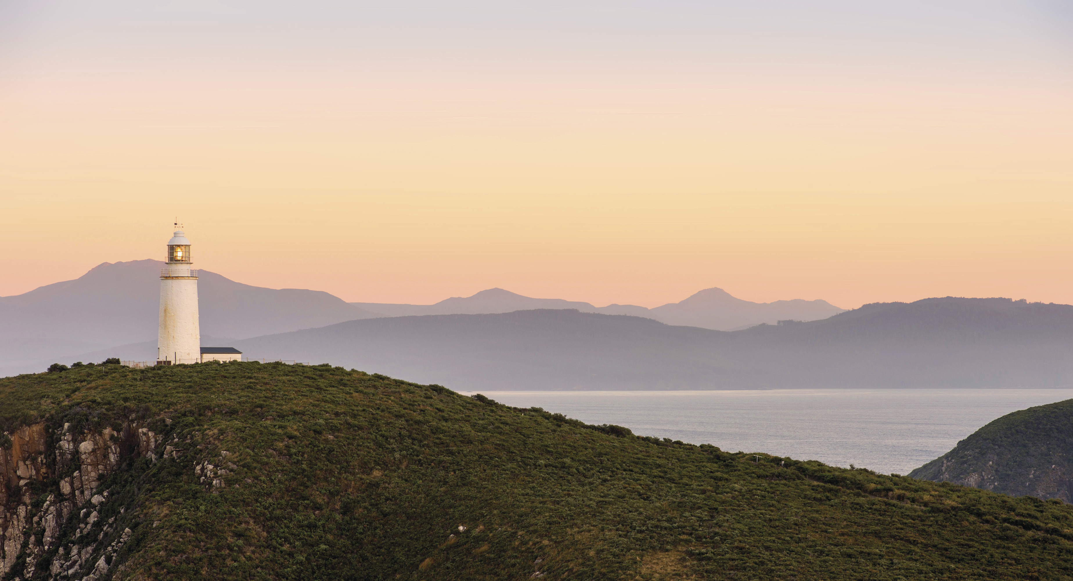 Stunning landscape image of Cape Bruny Lighthouse on sunset, with the ocean and mountains lie in the background.
