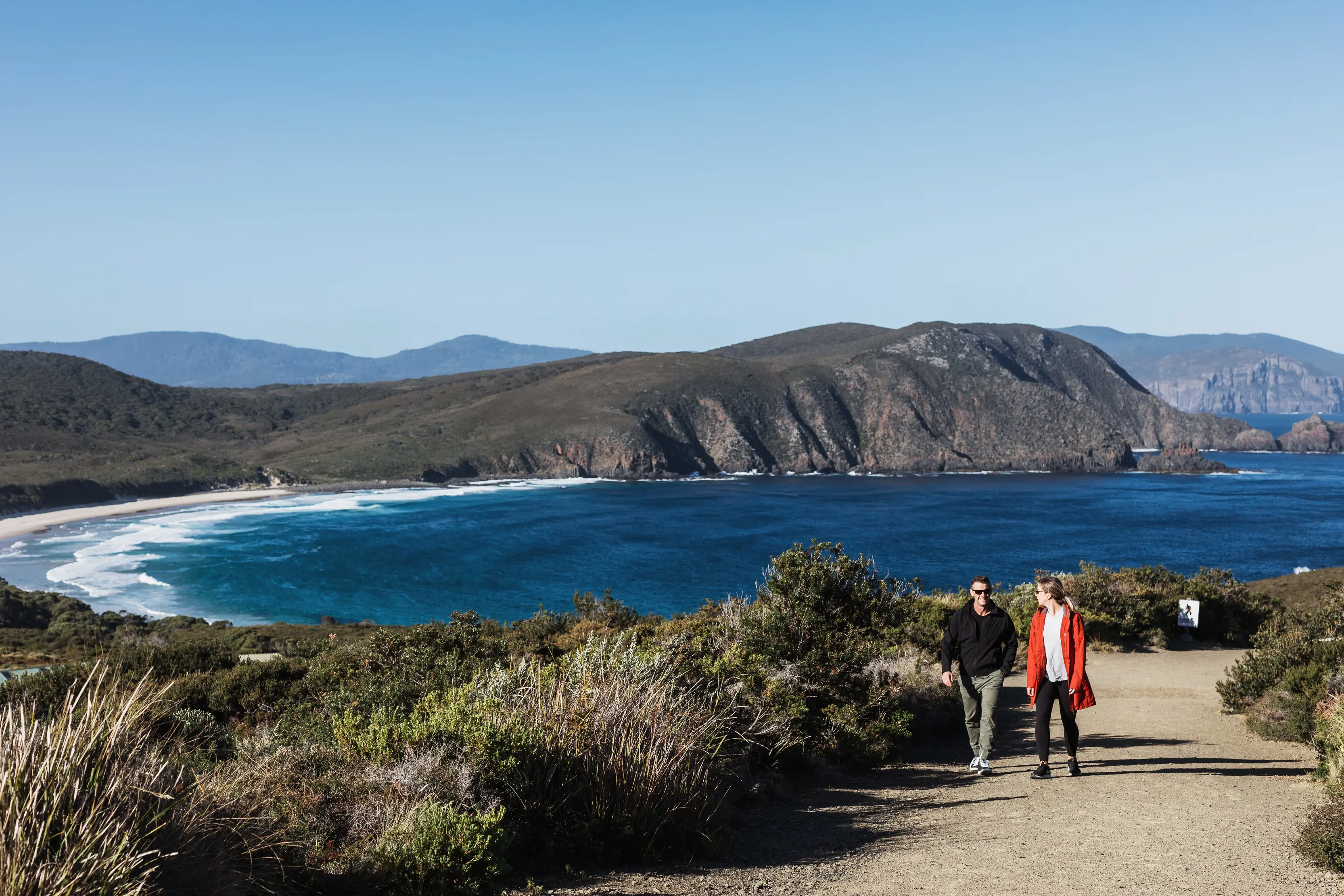 Couple walking up from Lighthouse Bay - Bruny Island on a pristine, sunny day, with striking blue water and lush bushland.