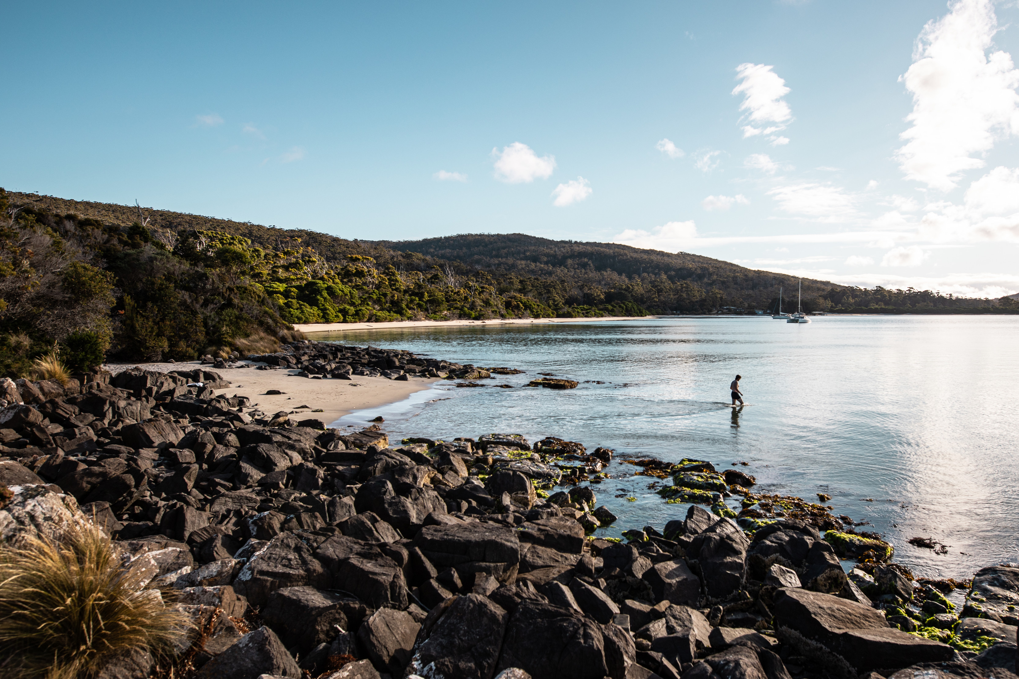 A man walks in the shallow water at Cockle Creek, the green landscape is in the background and rocks in the foreground.