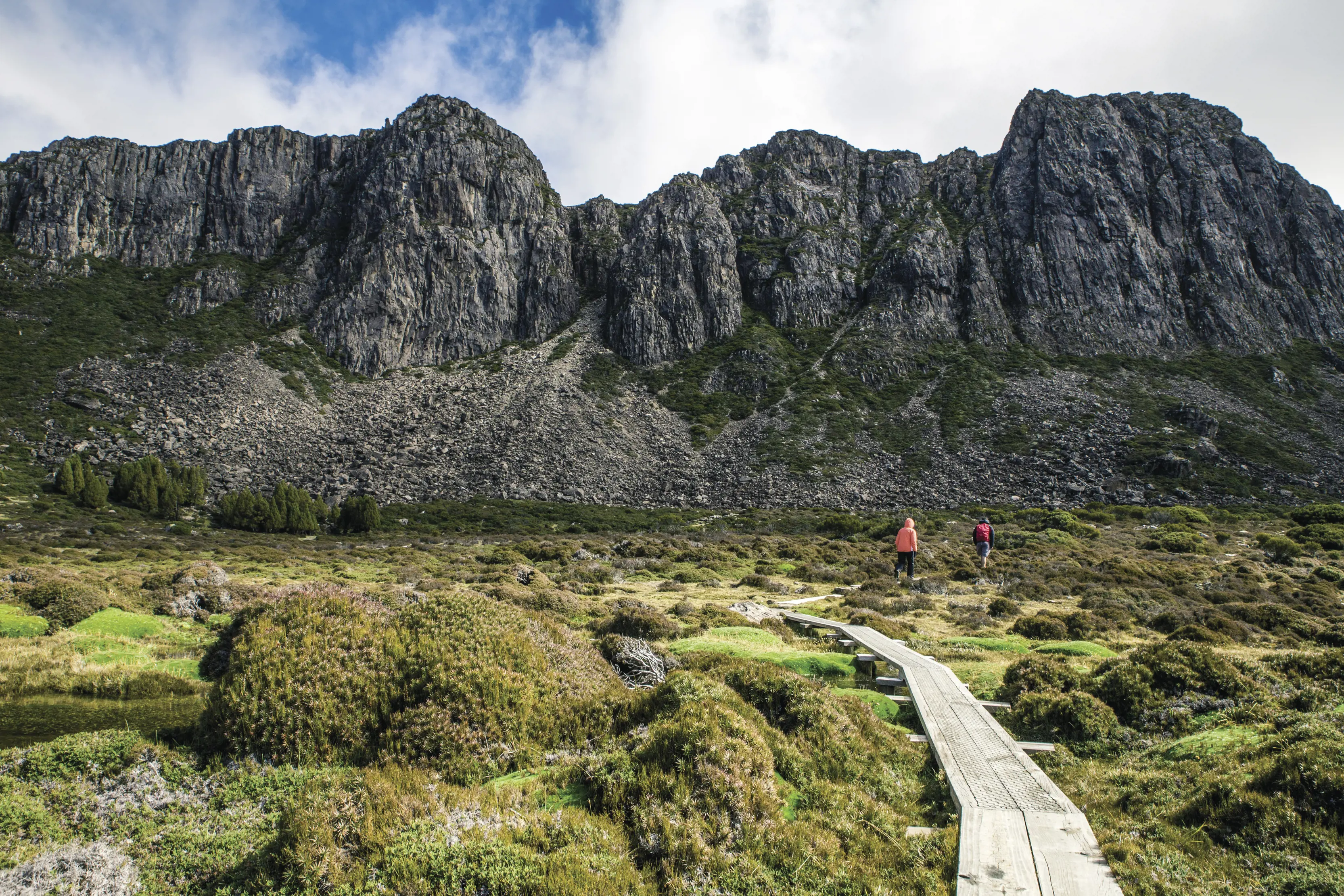 "Landscape image of two people walking on a footpath around the Walls of Jerusalem National Park, surrounded by large stone walls and lush bushland. "