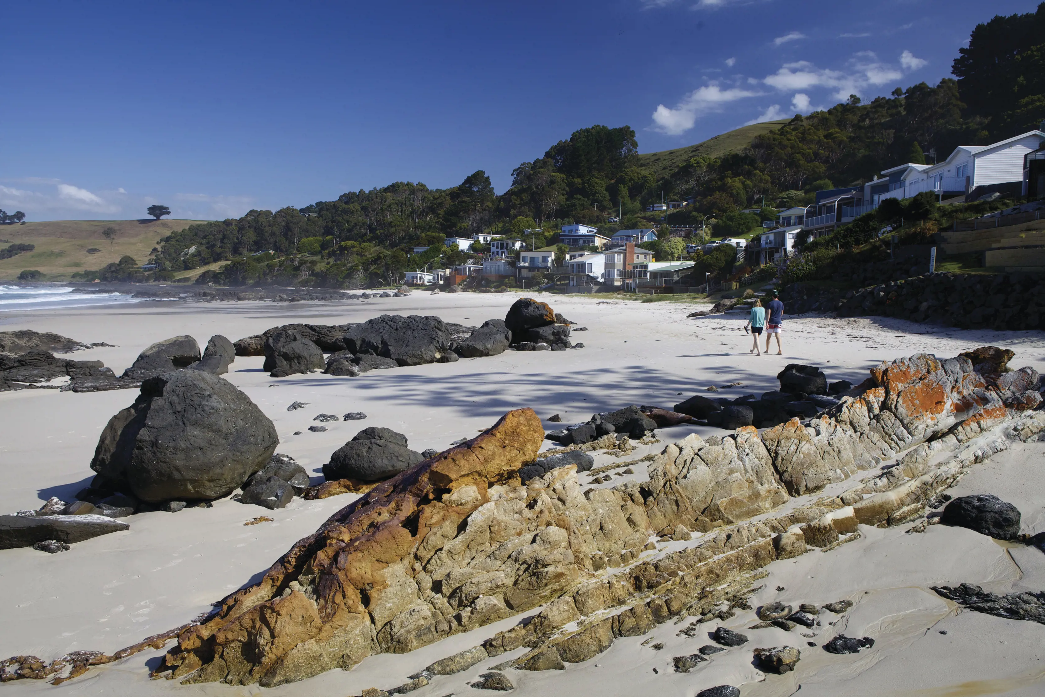 Two people walking on the sand, along the shore of Boat Harbour. Rocks fill the foreground of the image.