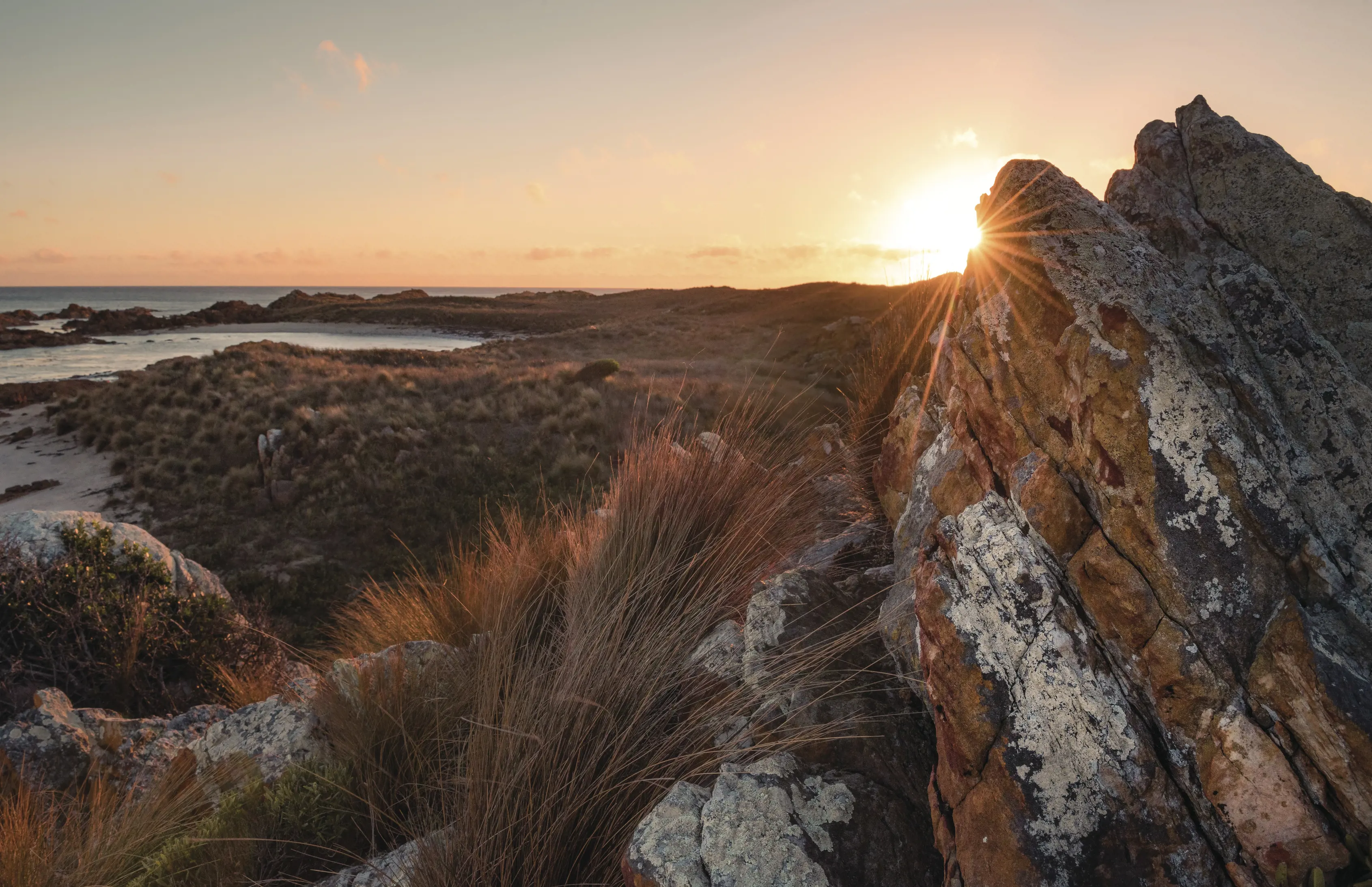 A stunning sunrise fills the Arthur-Pieman Conservation Area. The sun hits the surrounding bushland and rocks in the image.