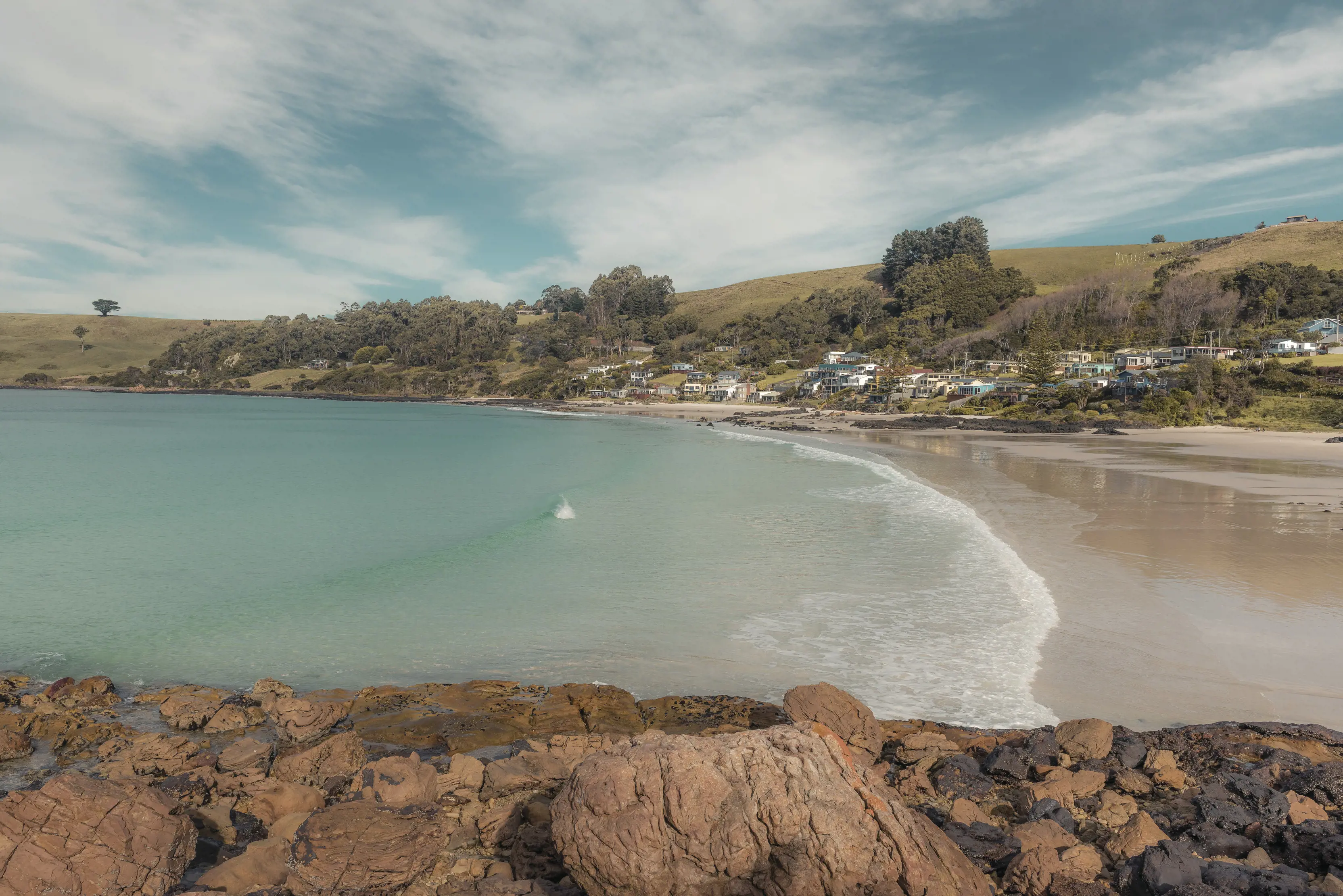 Picturesque view of Boat Harbour from a side angle. The sand meets the clear blue water and the houses and bushland are in the background.  