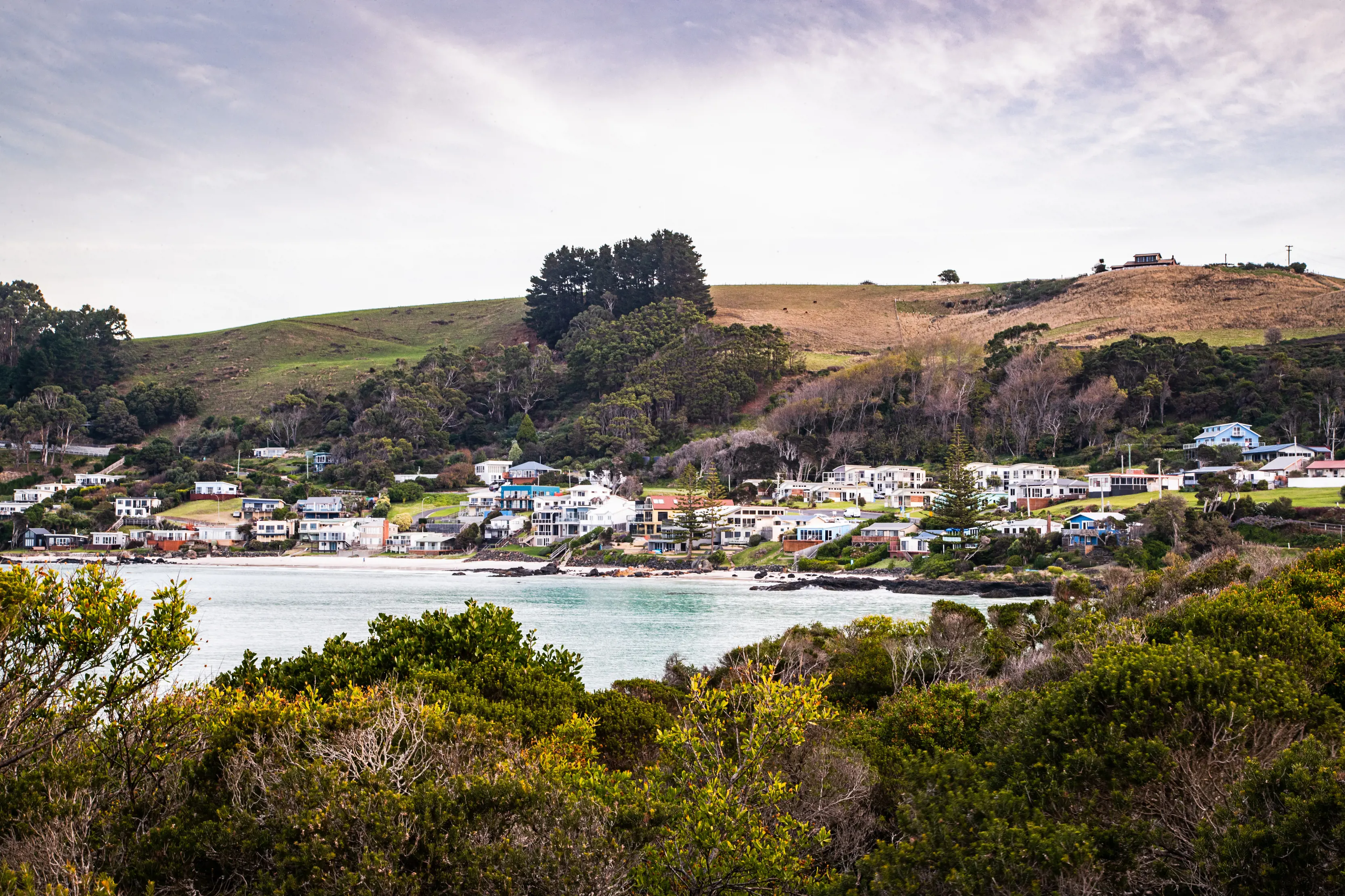 Bushes fill the foreground of the image with the ocean and houses of Boat Harbour surround the centre of the image. Valleys fill the background of the image.