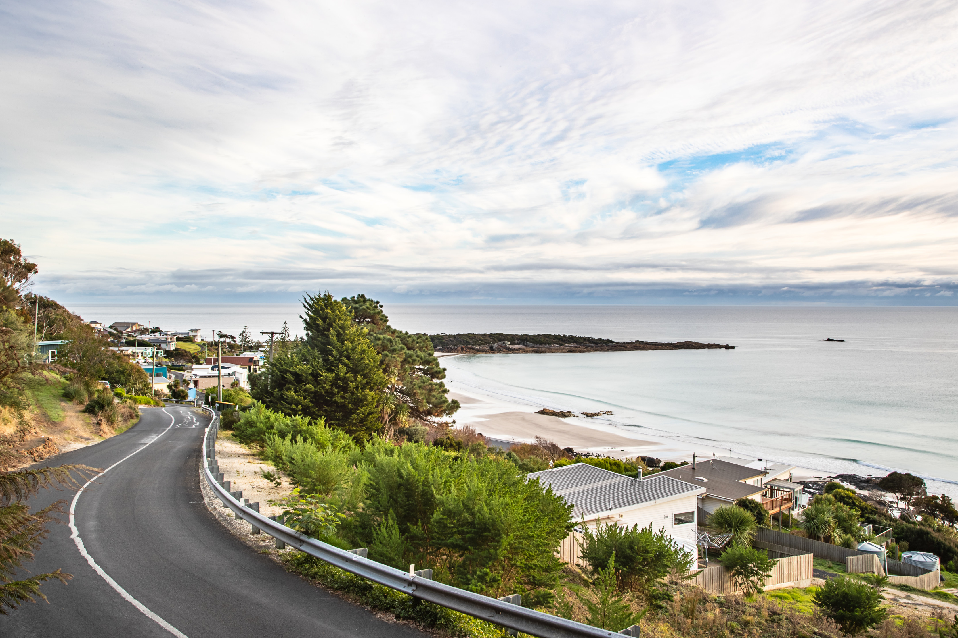 Image of a windy road into Boat Harbour on the left side of the image. Houses fill the right of the image that lead to the shoreline and ocean. 