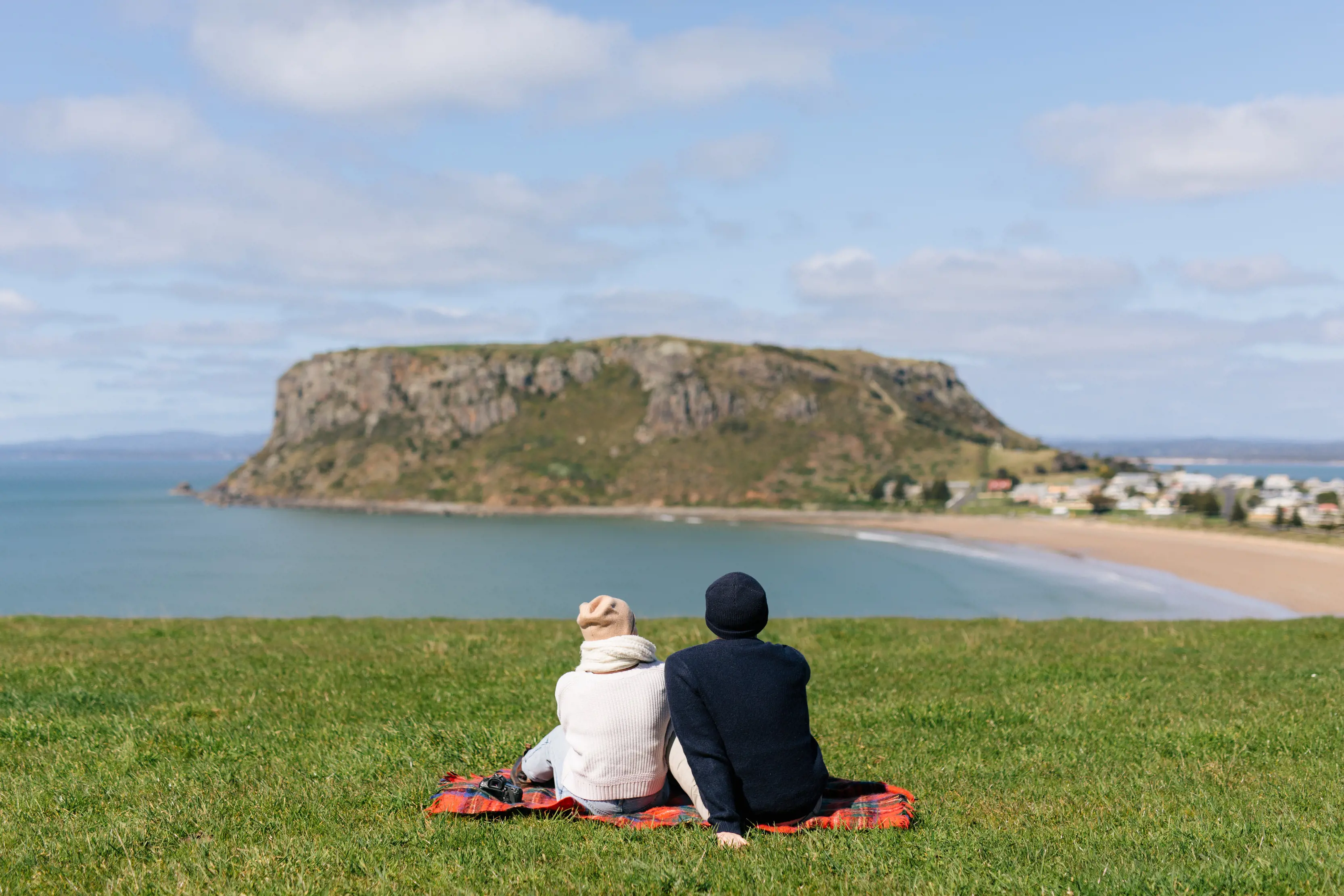 A couple, positioned in the centre of the image, sit on a hill, overlooking The Nut and the village of Stanley.