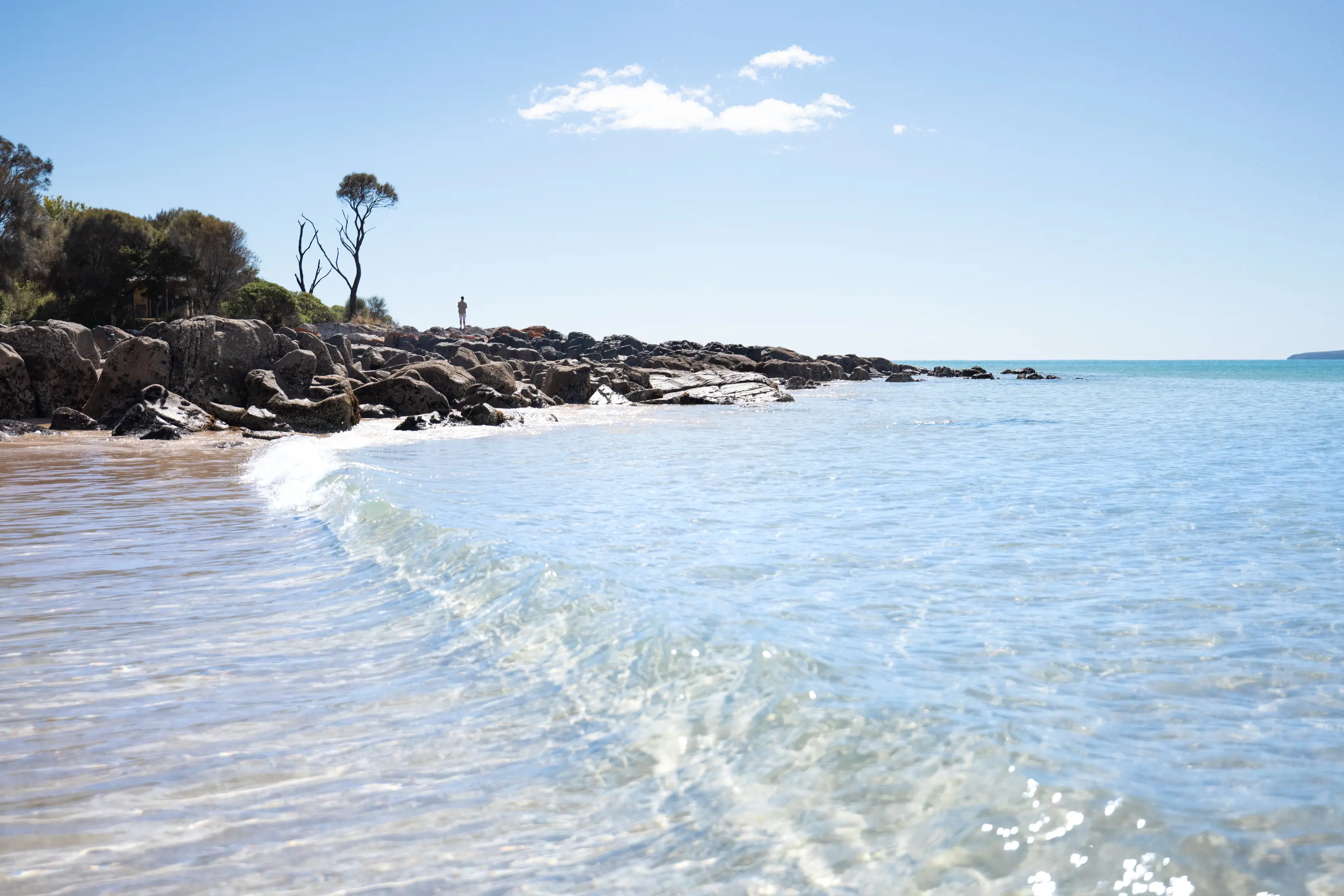 Image of the pristine water, on the shore of Hawley Beach, with a human on the rock point in the background, on a clear, sunny day.