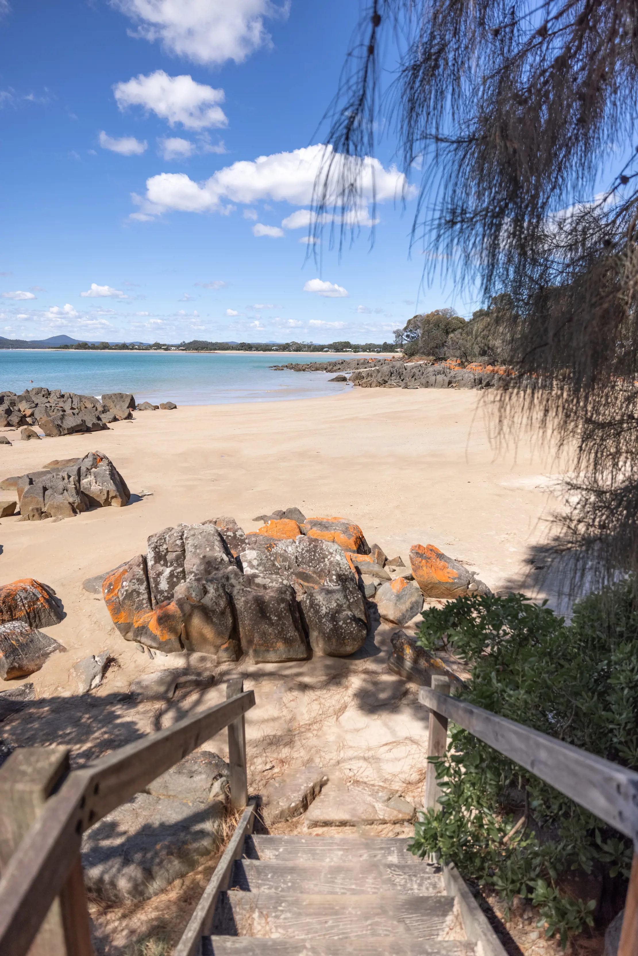 Wooden stairs at the foreground of the image, leading to the sand of Hawley Beach, with rocks amongst the sand and the ocean in the background. 