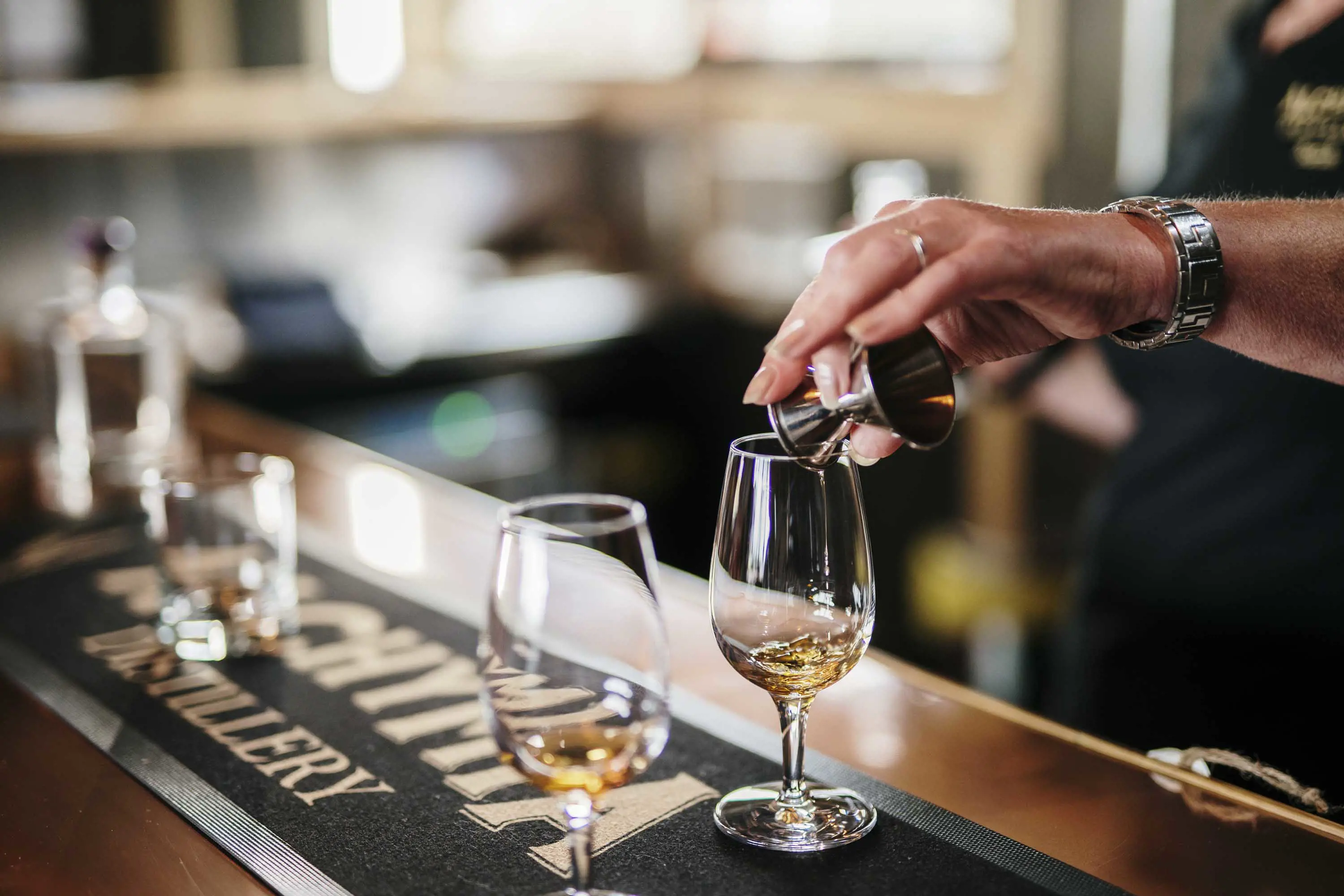 A bartender pours a dram of whisky from a metal shot glass into a tasting glass.