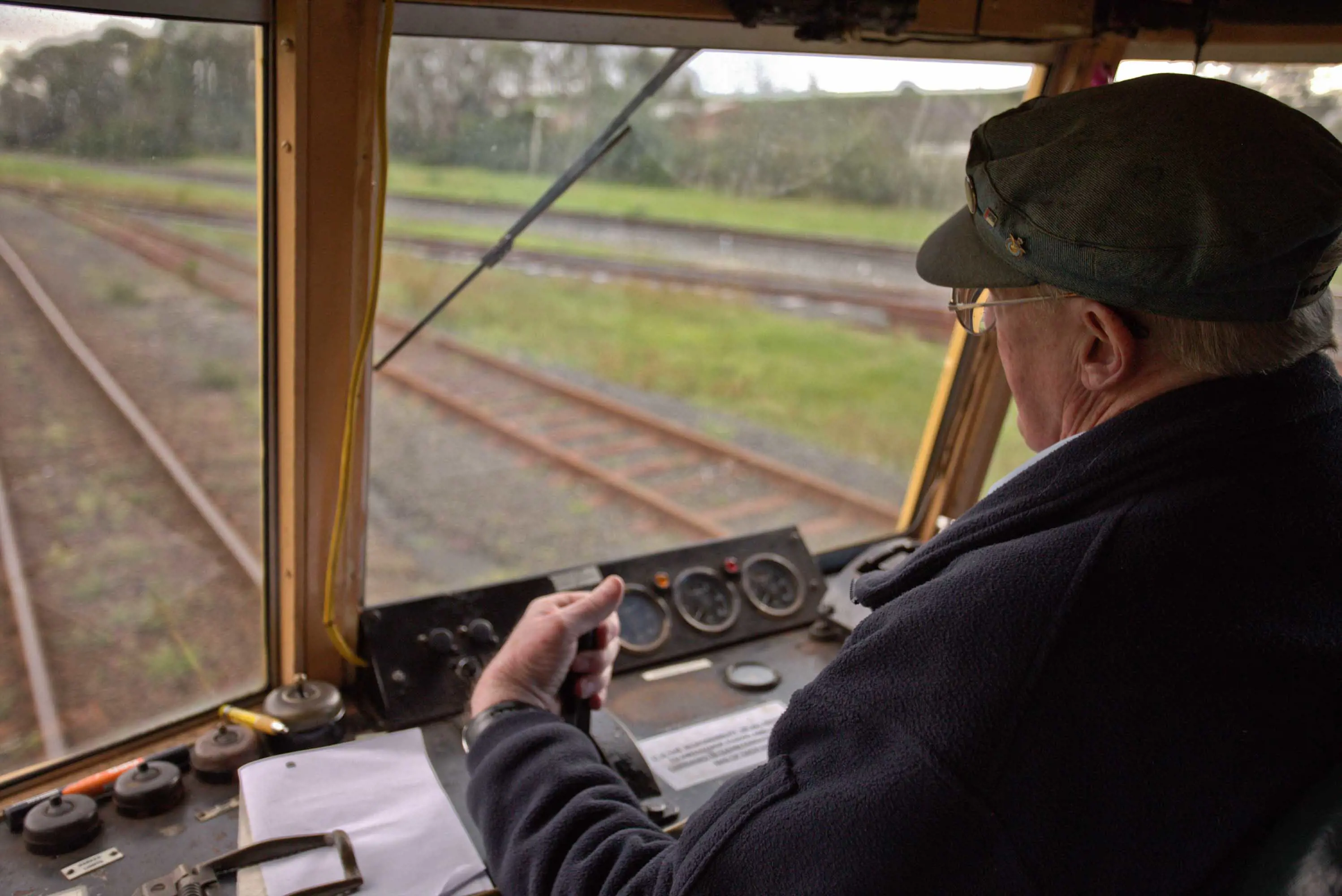 A man grips a lever on a panel of controls at the front of an old train, which is travelling along one of a series of railway lines.  
