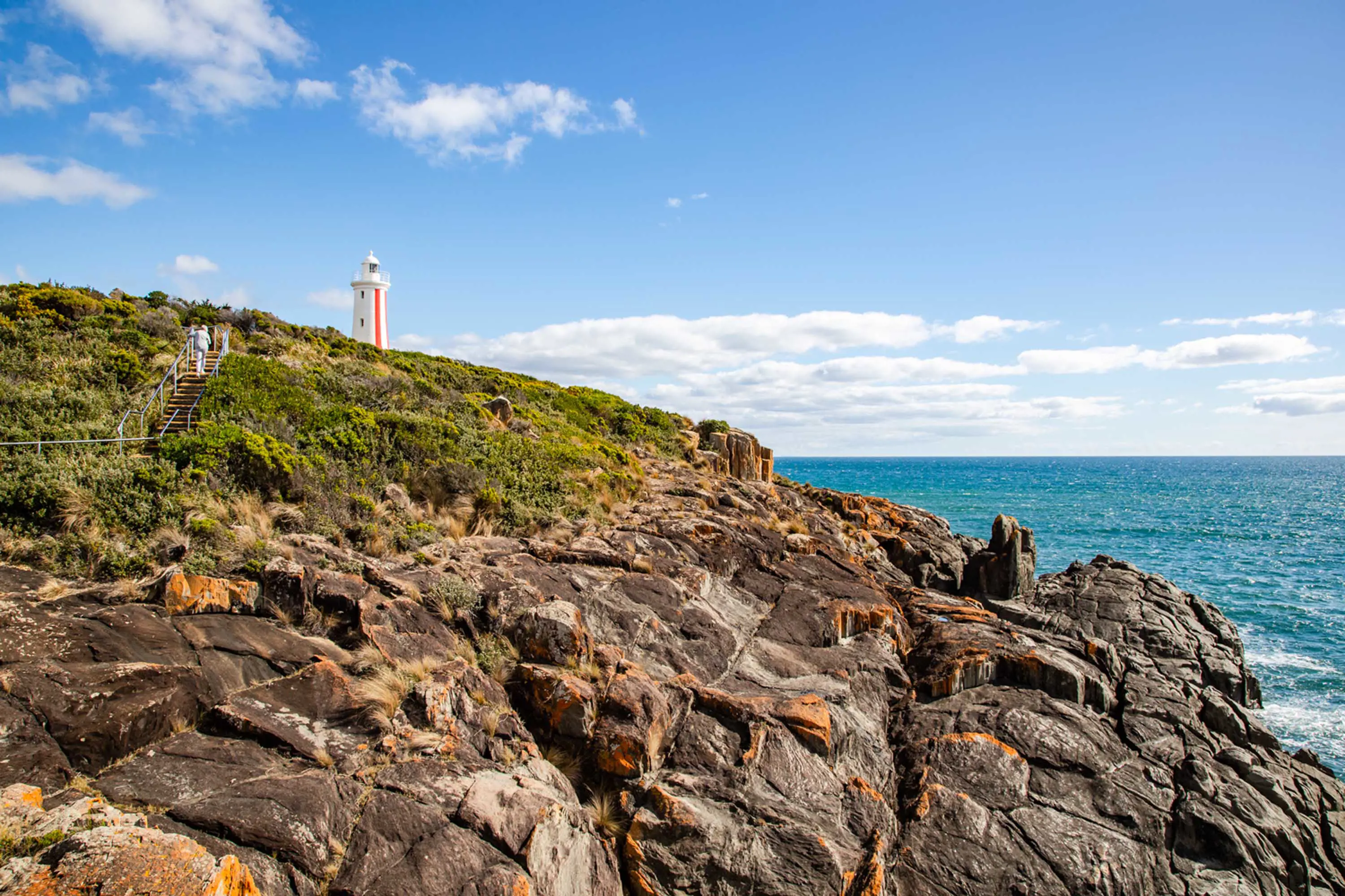 Oramge lichen covered rocks of a point looking out over water. A white lighthouse with a red stripe up one side stands in the distance.