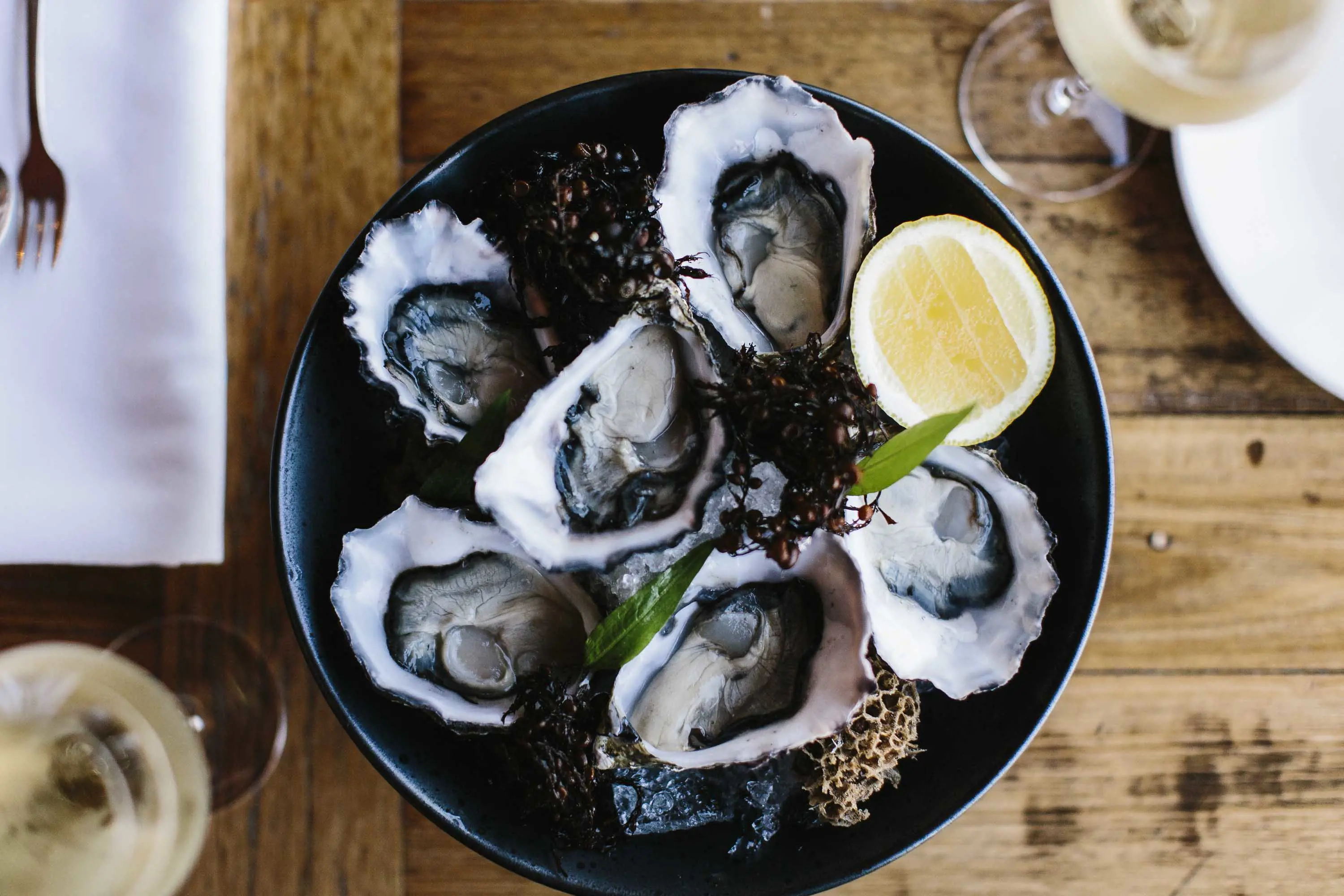 A plate of fresh oysters with garnish and a slice of lemon, presented on a black plate sitting on a wooden table.