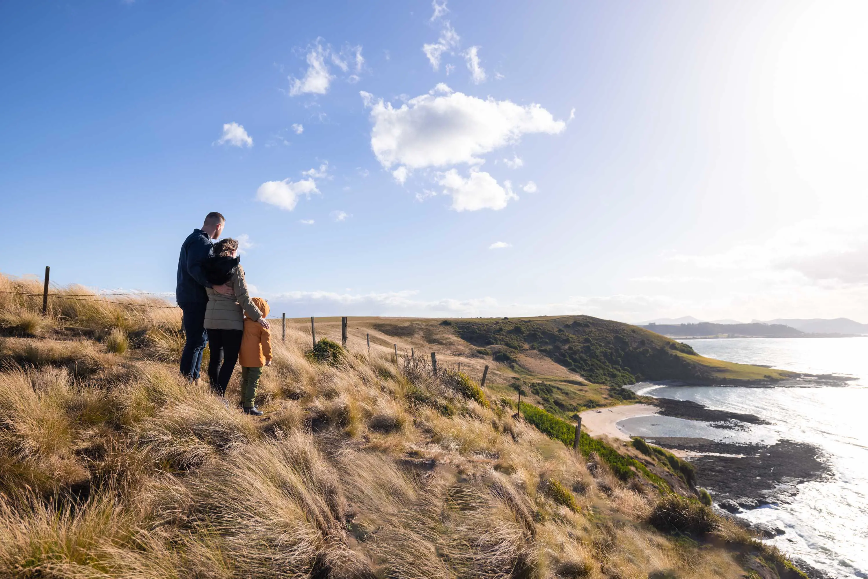 A young family wearing wet-weather jackets stand together on a grassy hill looking down a rocky coastline on a sunny day.