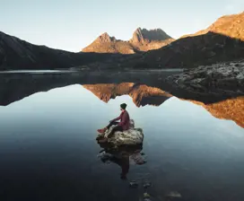 Person sitting on a rock in the middle of a lake amongst Cradle Mountain. The jagged contours of Cradle Mountain surround the image.