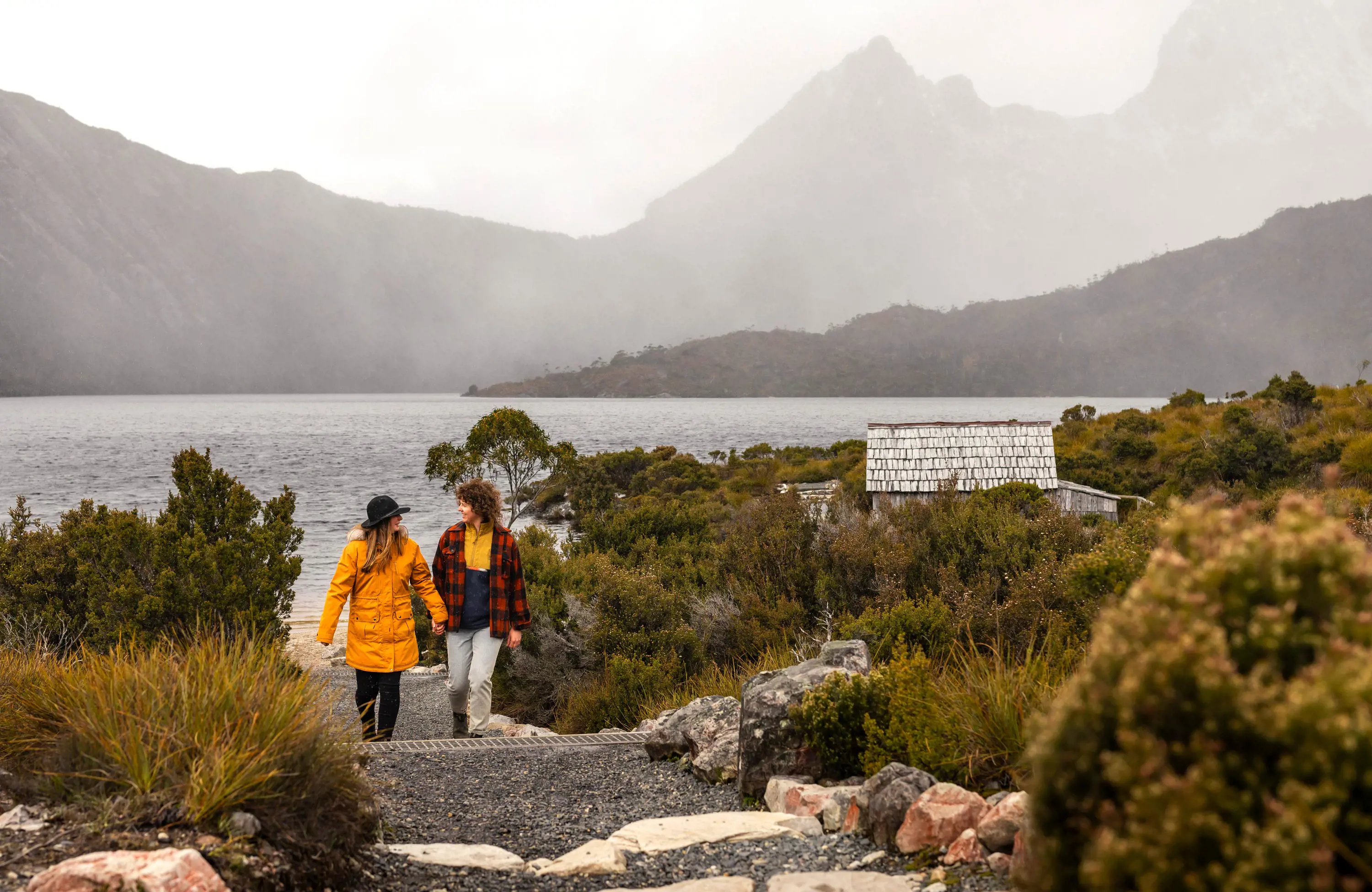 A young couple walk up a gravel path beside Dove Lake at Cradle Mountain on a misty day.