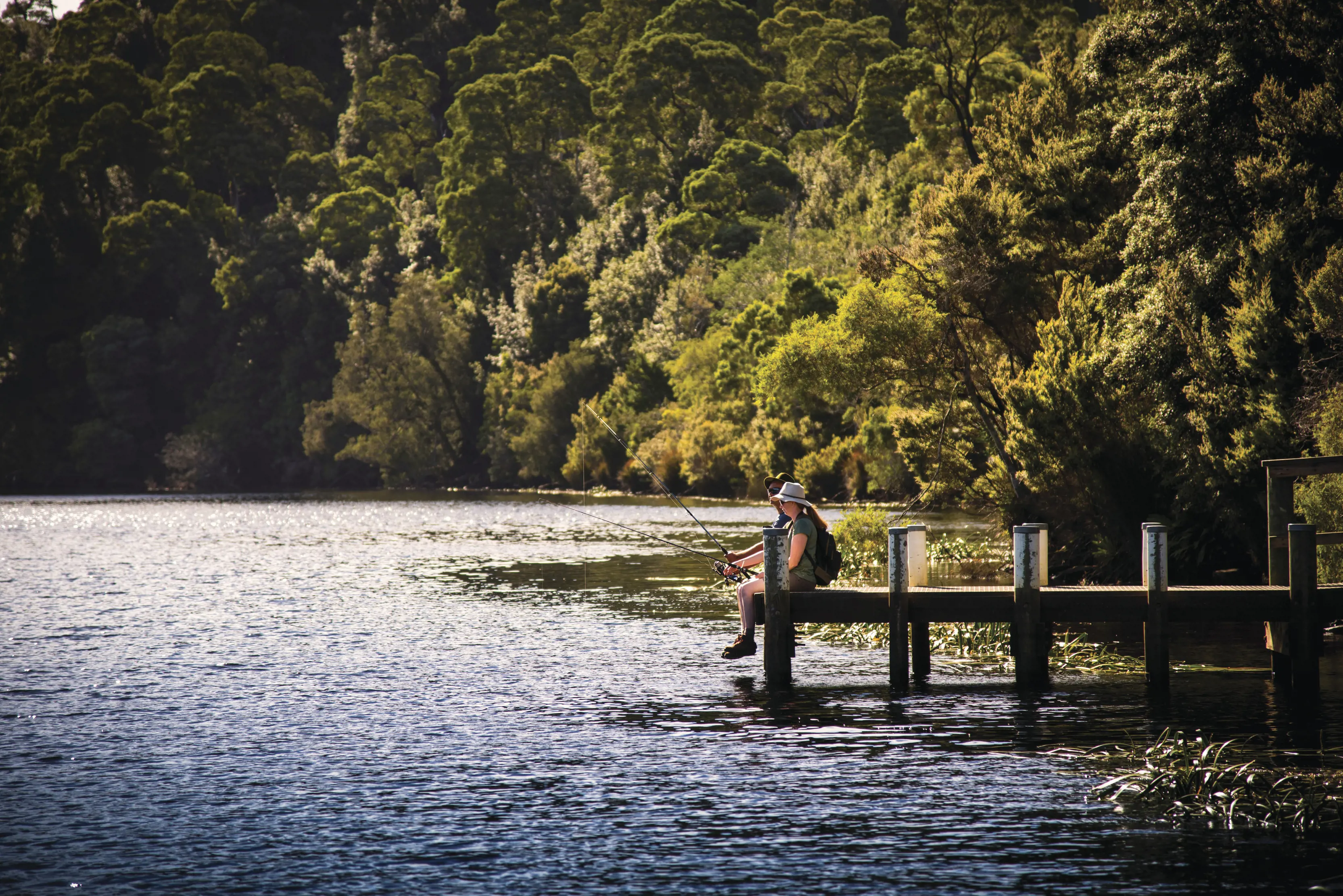 Woman sits on the end of a jetty on a beautiful summer's day, fishing on the Pieman River.