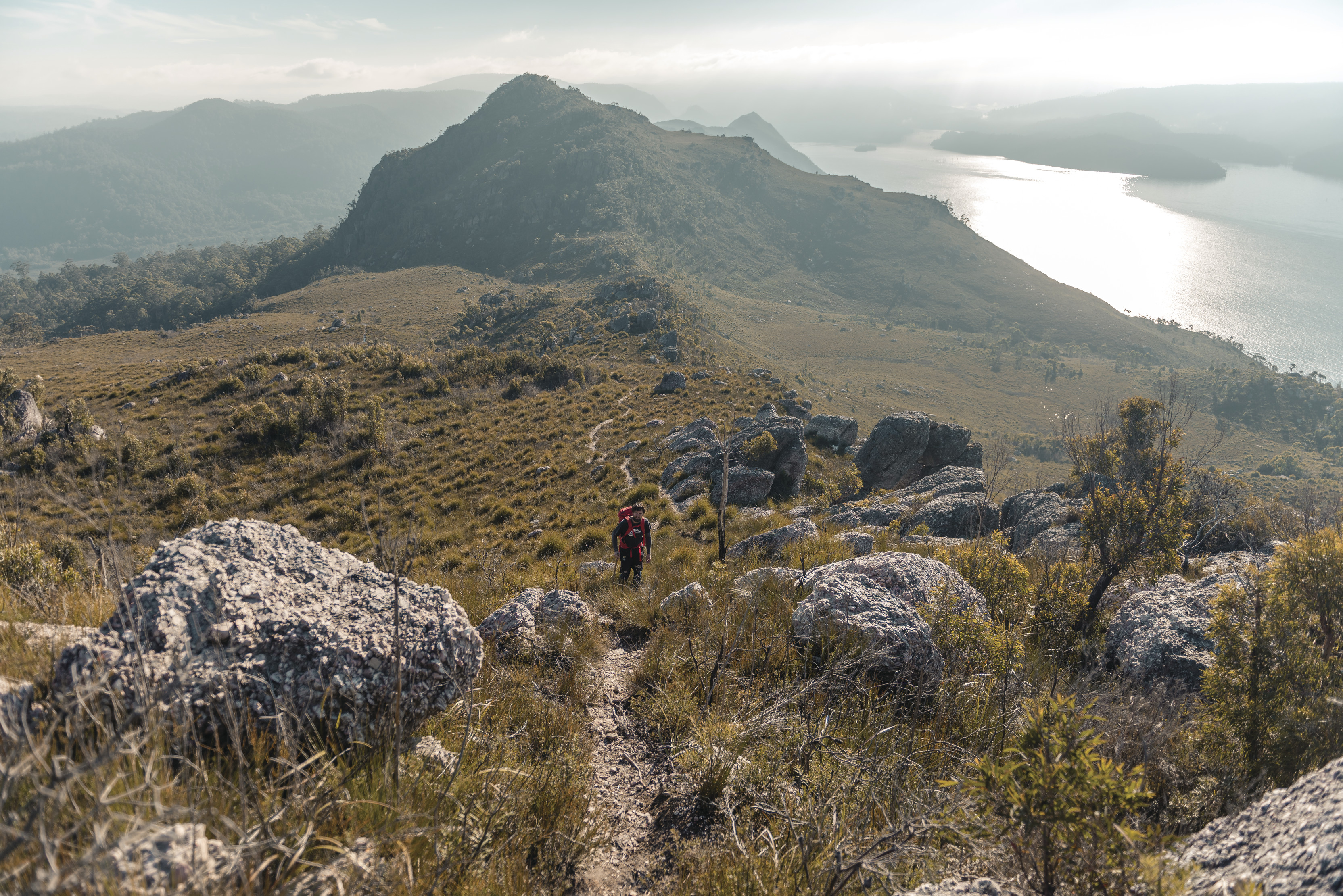 A single hiker walks along the track at Mt Farrell, water and mountain views in the background.