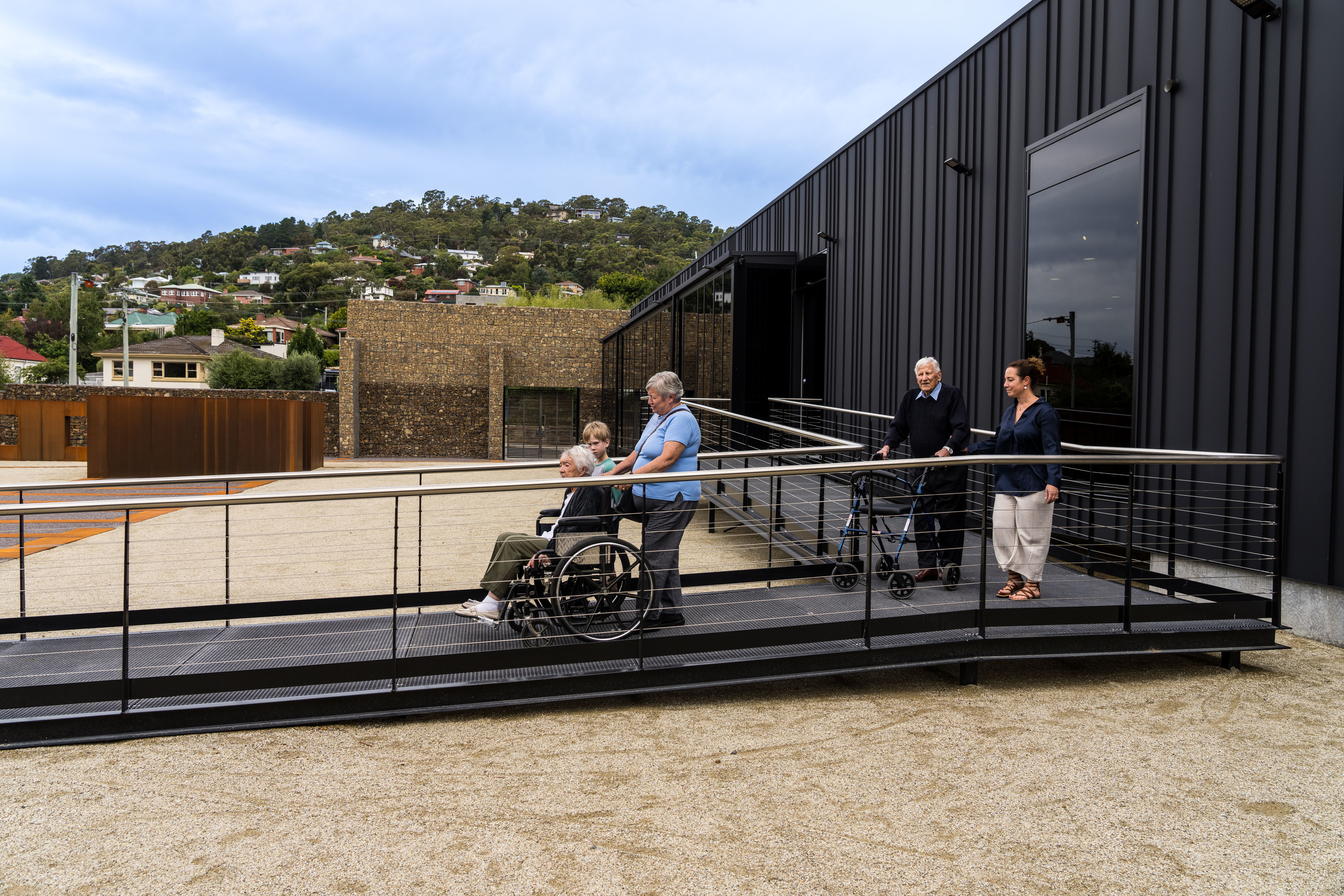 Two groups of people outside the Cascade Female Factory Historic Site, one being in a wheelchair and one with a walking frame, walking down the wheelchair access ramp.