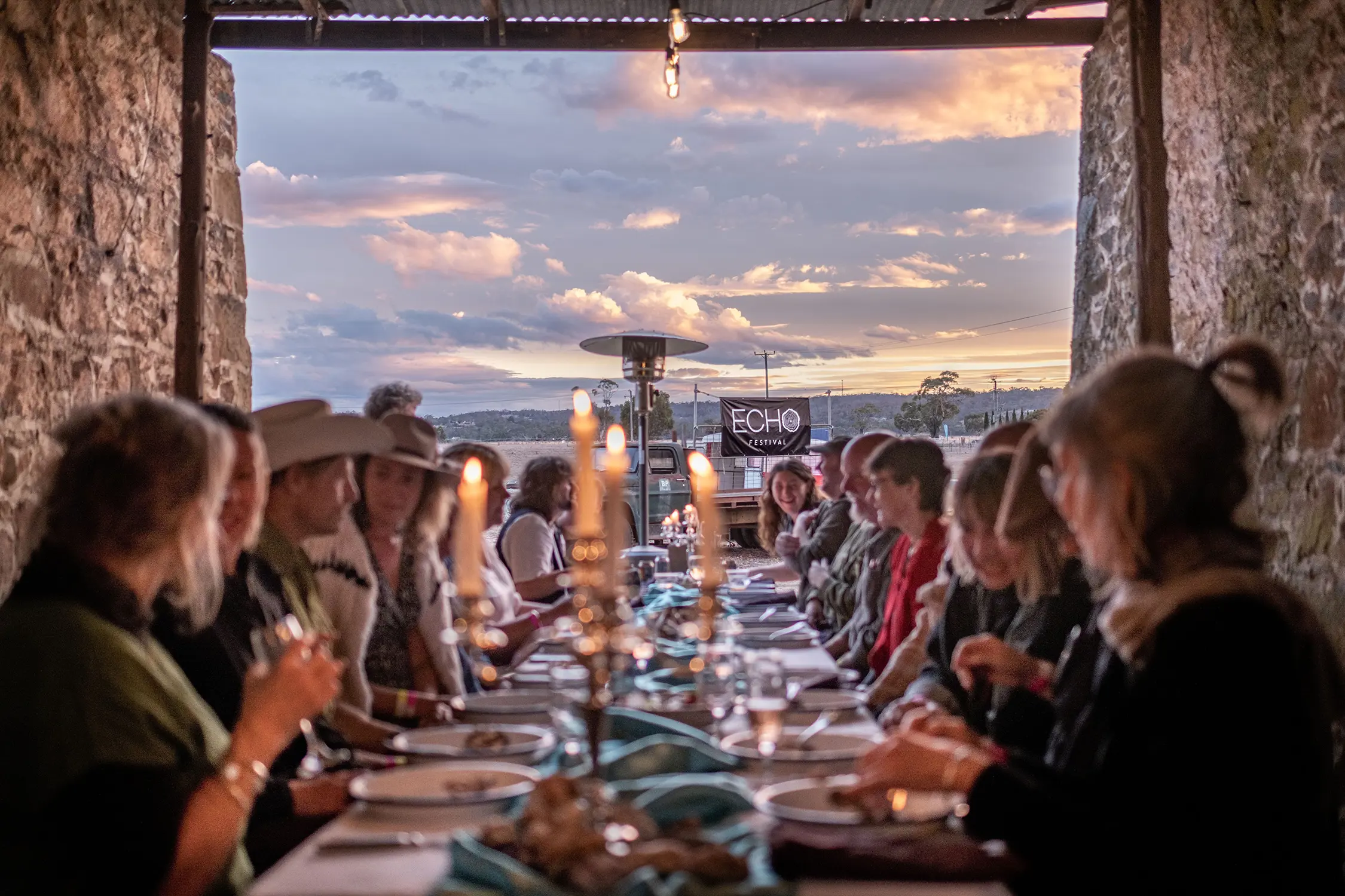 Looking down a long table with people seated either side, eating plates of food and chatting. The sunset colours clouds in the background.