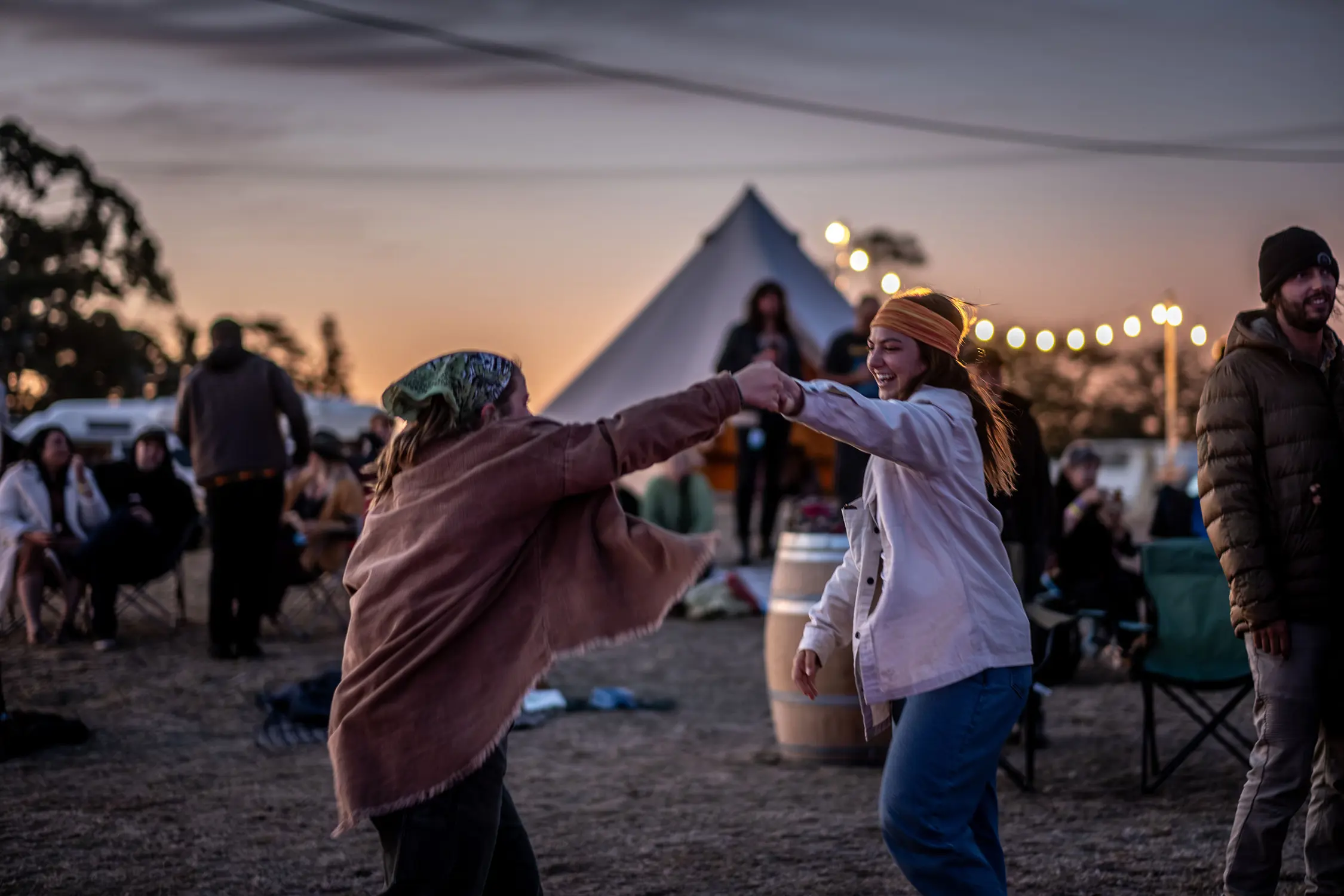 At an outdoor music festival at dusk, two people hold hands and dance, surrounded by tents, fairy lights and other festival-goers.